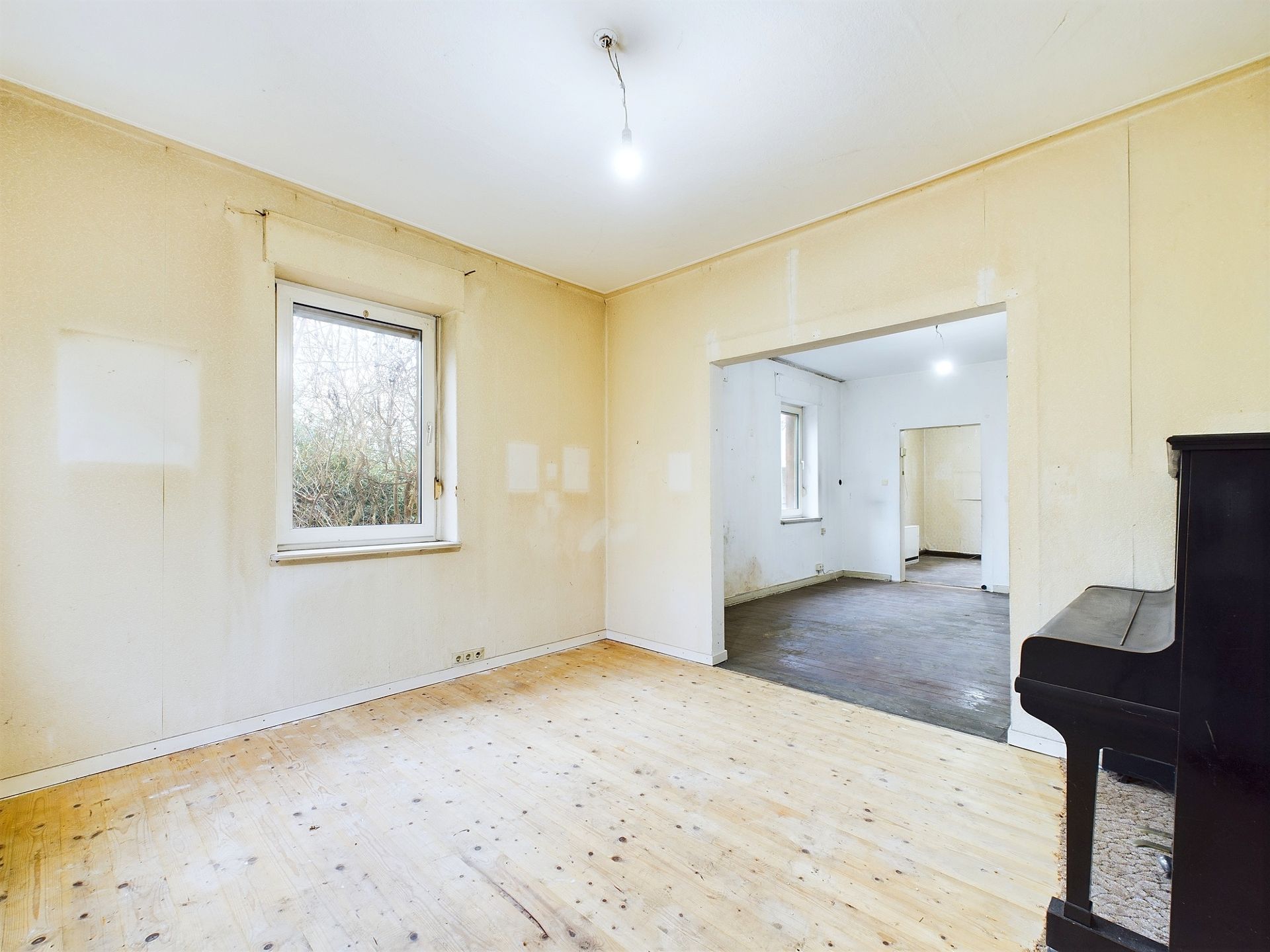 Heller Raum mit Holzfußboden, Fenster und einem dunklen Klavier, angrenzend an weiteren Raum.