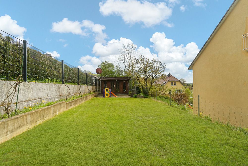 Gepflegter Garten mit Spielhaus und Spielgeräten, umgeben von Zaun und Wohnhäusern.