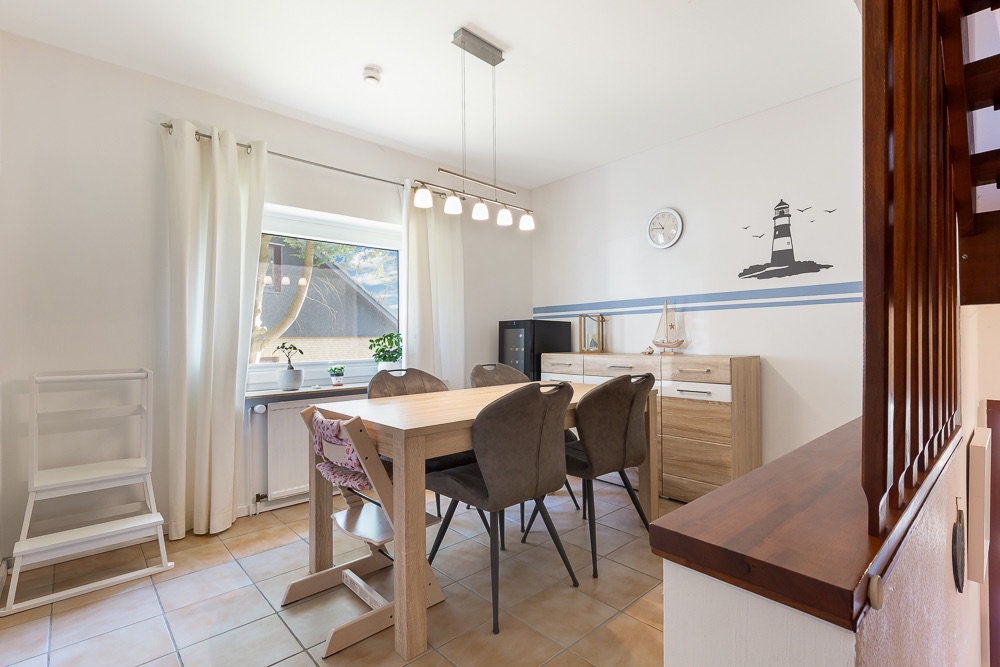 Esszimmer mit Holztisch, sechs Stühlen, Sideboard, Wandbild eines Leuchtturms und großem Fenster mit weißen Vorhängen.