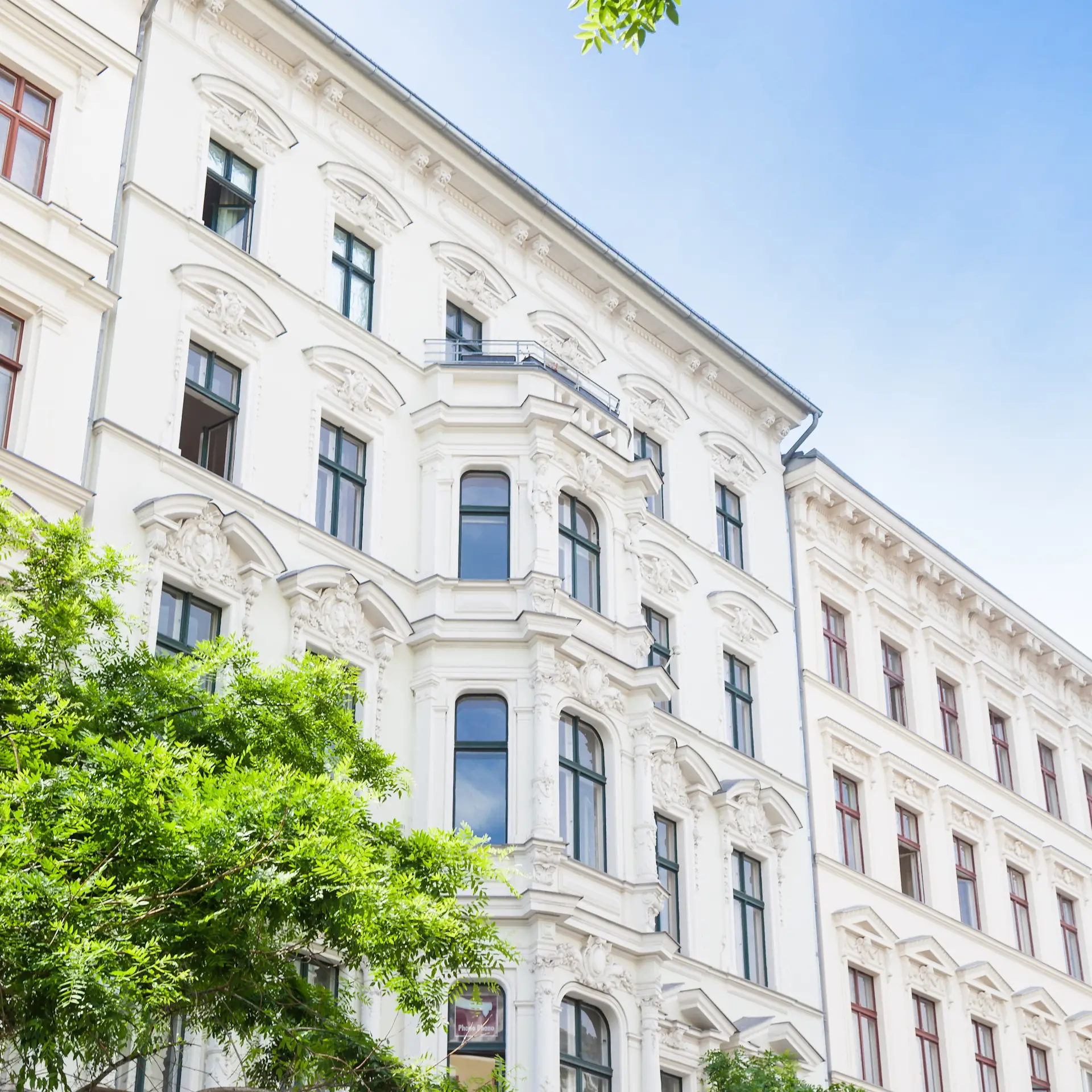 Fassade eines hellen Wohnhauses mit Stuckverzierungen und großen Fenstern, Bäume im Vordergrund.