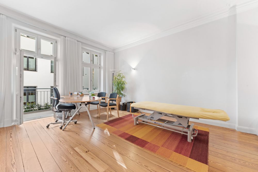 Heller Raum mit Holzboden, Tisch, Stühlen, gelber Liege und großem Fenster.