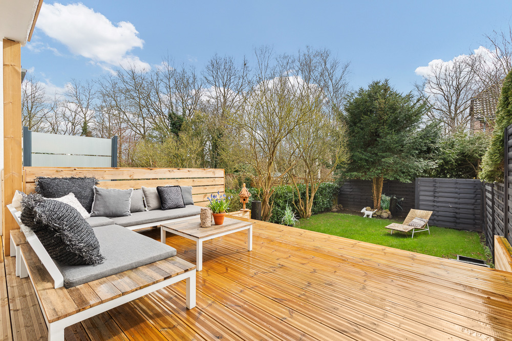 Holzterrasse mit Sitzbank und Tisch, Blick auf Garten mit Liege und Bäumen.