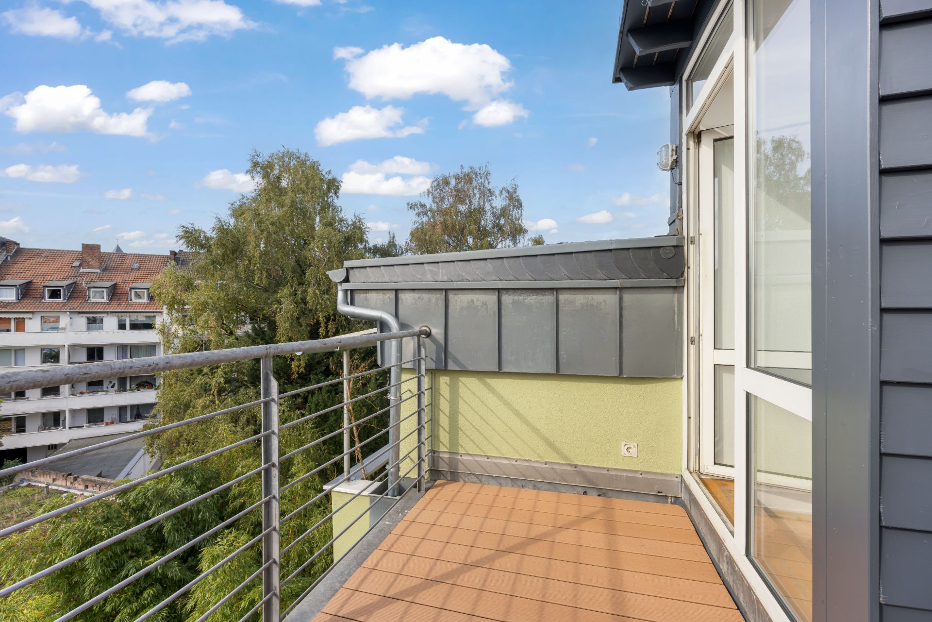 Kleiner Balkon mit Metallgeländer, Blick auf Bäume und Nachbarhäuser, offene Glastür.