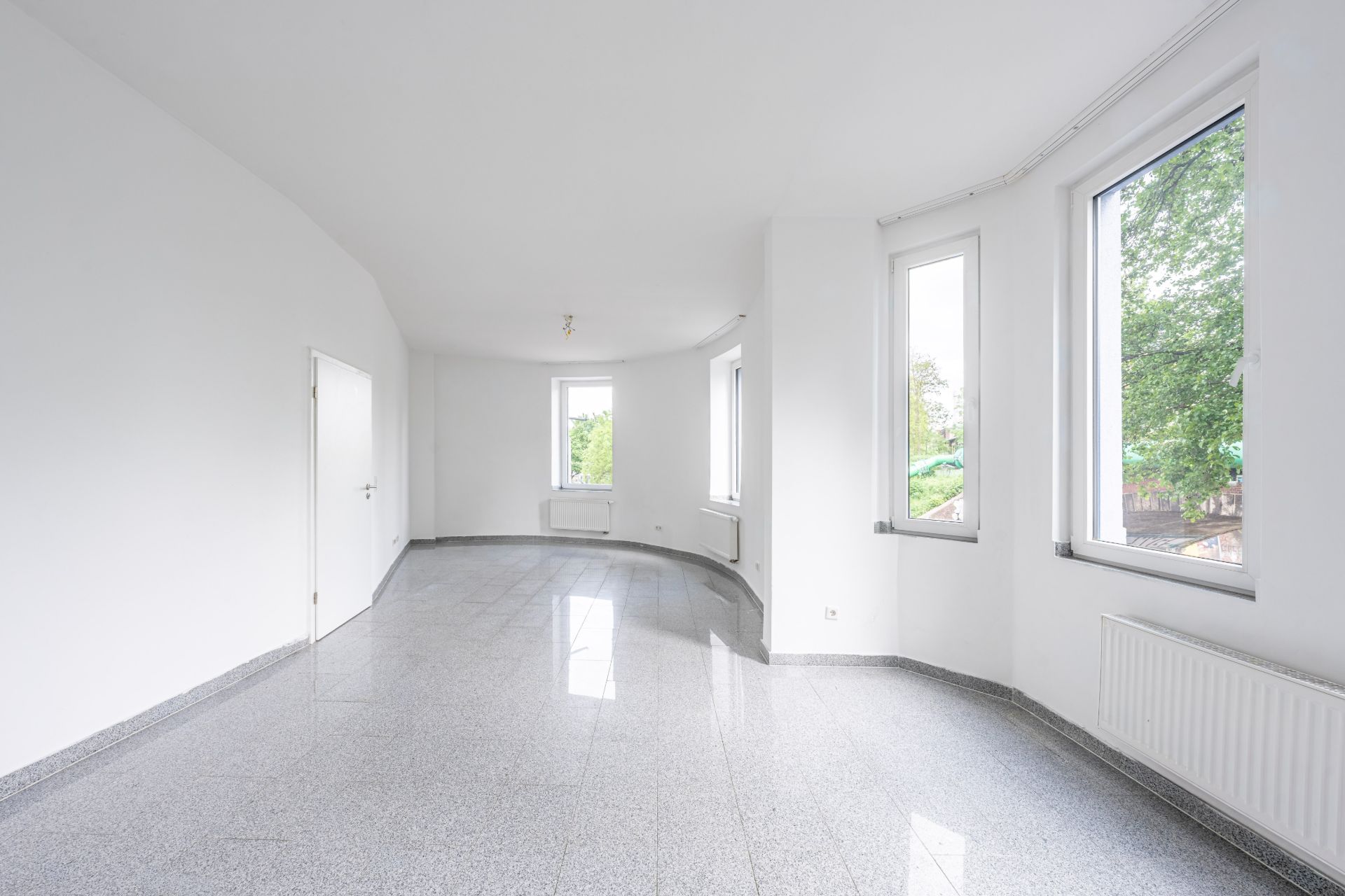 Heller leerer Raum mit großen Fenstern, weißen Wänden und grauem Boden.