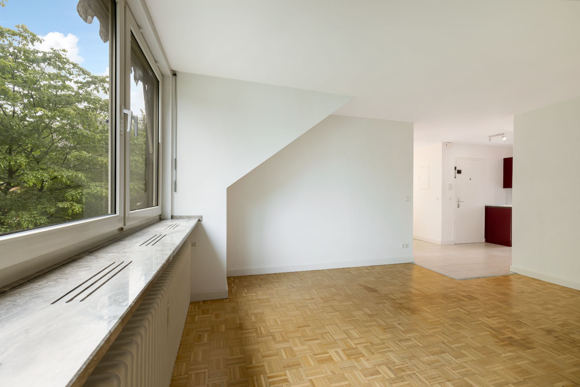 Helles Wohnzimmer mit Parkettboden, großem Fenster und Blick auf Bäume, offene Verbindung zur Küche.