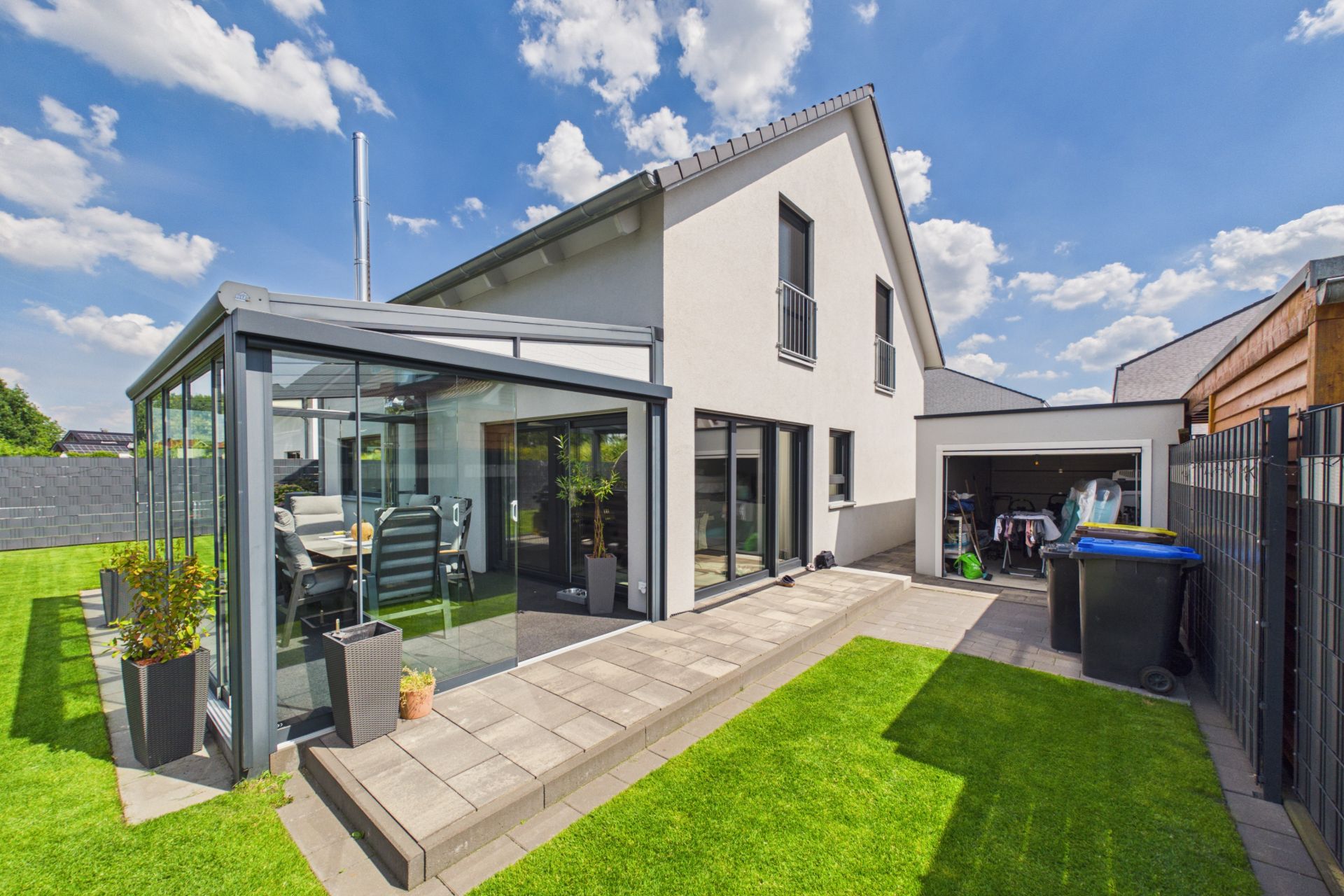 Modernes Wohnhaus mit Wintergarten, Terrasse, Garten und offenem Garagenbereich bei Tageslicht.