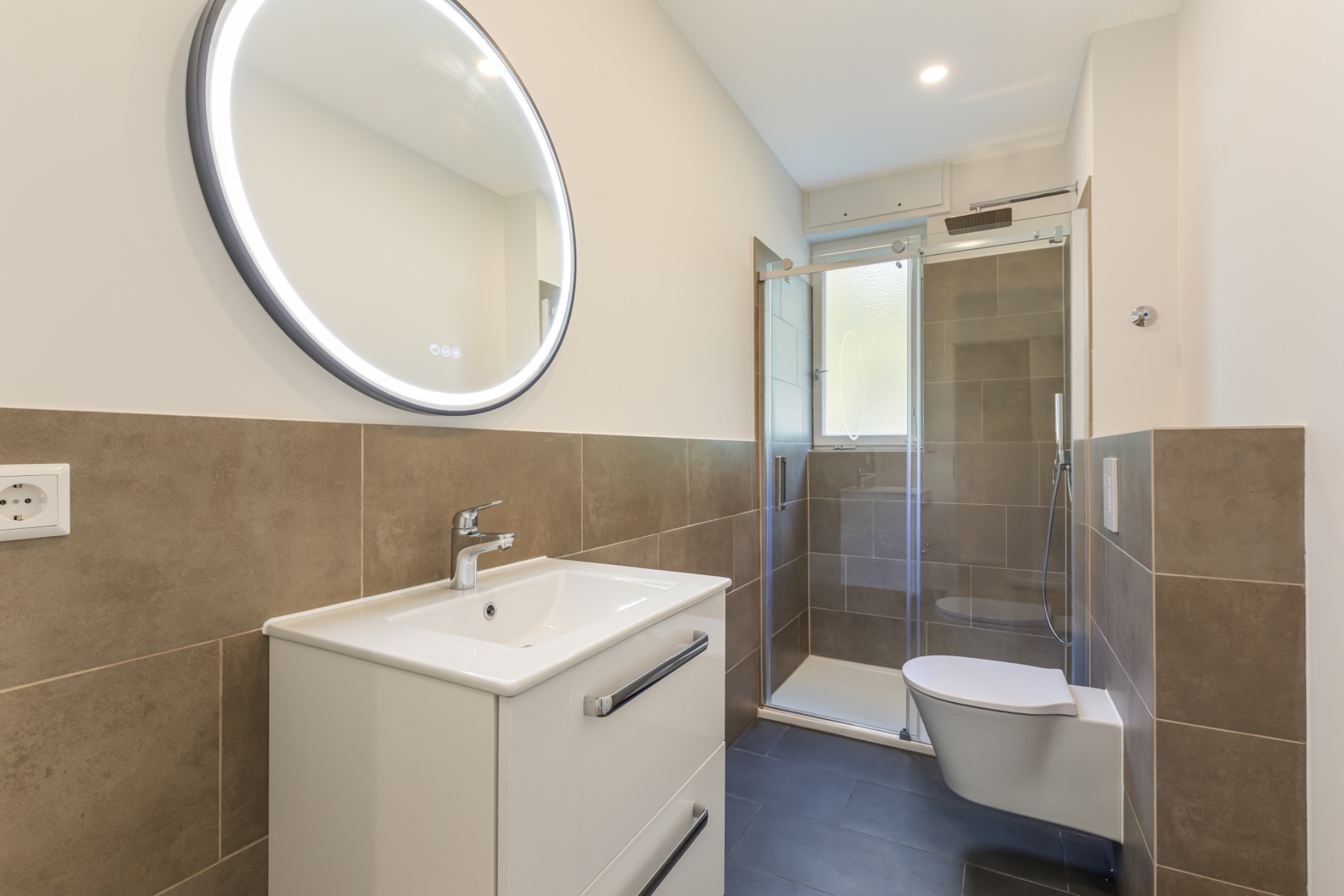Modernes Badezimmer mit beleuchtetem Spiegel, Waschbecken, Wandtoilette und Glasdusche.