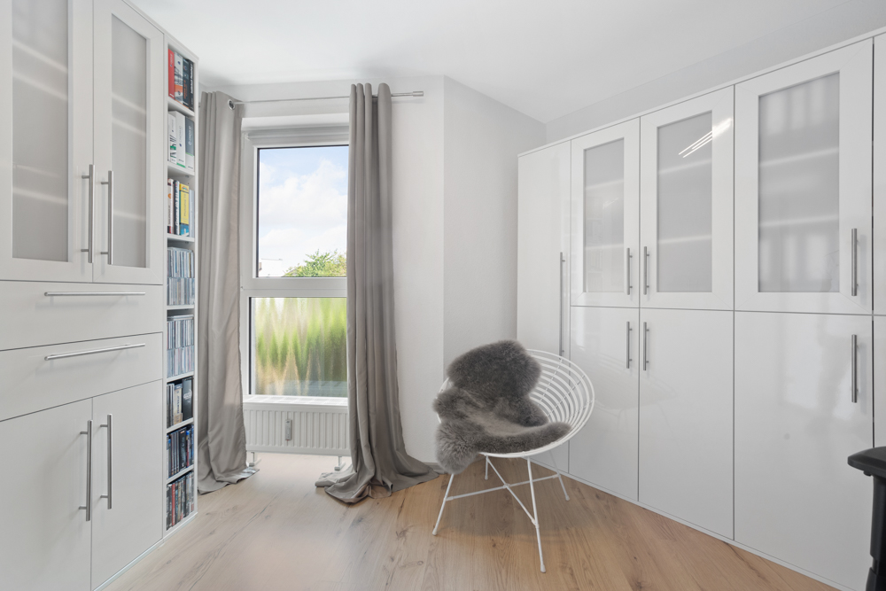 Heller Raum mit weißen Schränken, Bücherregal, Fenster und Stuhl mit Fellauflage.