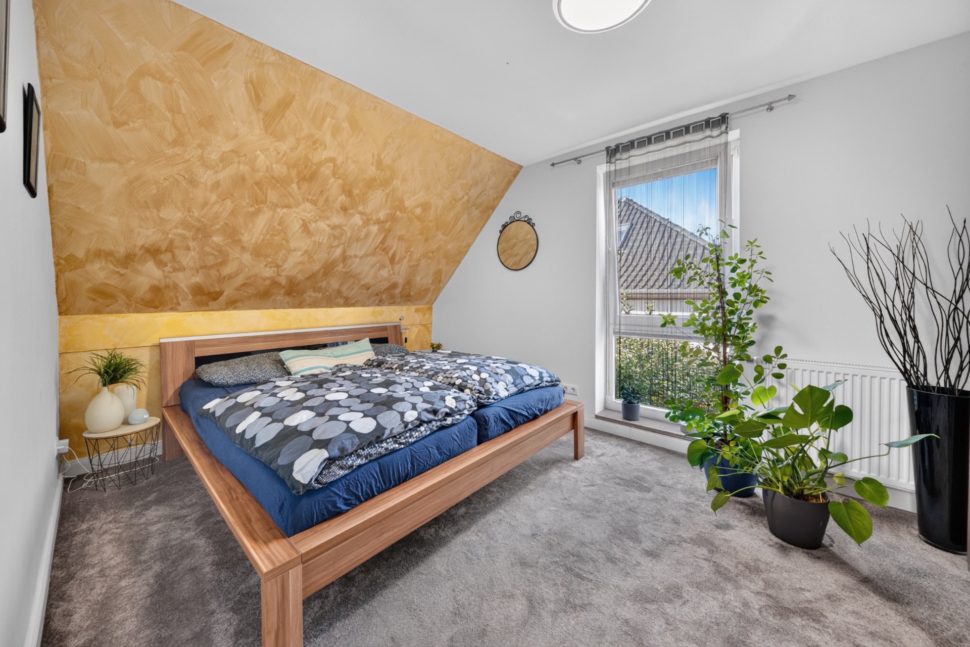 Schlafzimmer mit Bett, gelber Schräge, großem Fenster und Zimmerpflanzen.