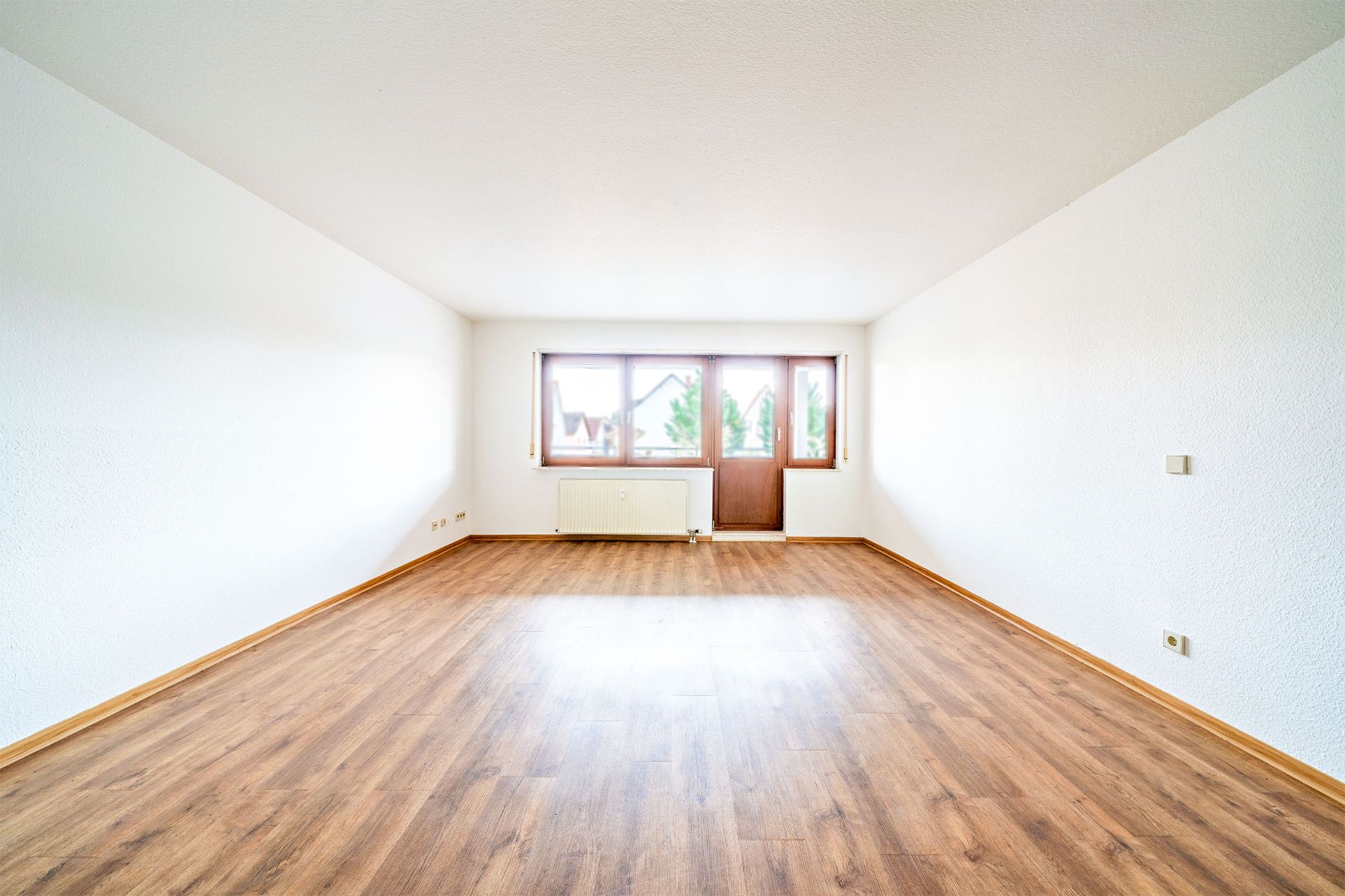 Leerer Raum mit Holzboden, weißen Wänden und großem Fenster mit Heizung.