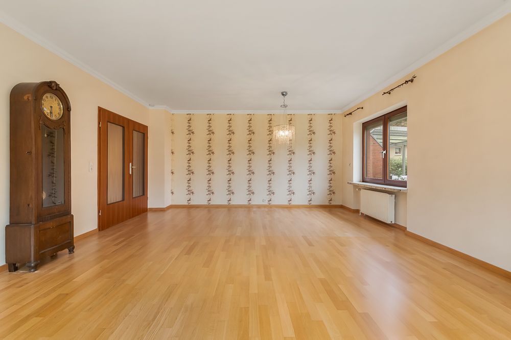Helles, leeres Wohnzimmer mit Holzfußboden, Standuhr, großem Fenster und Tapete mit Blumenmuster.