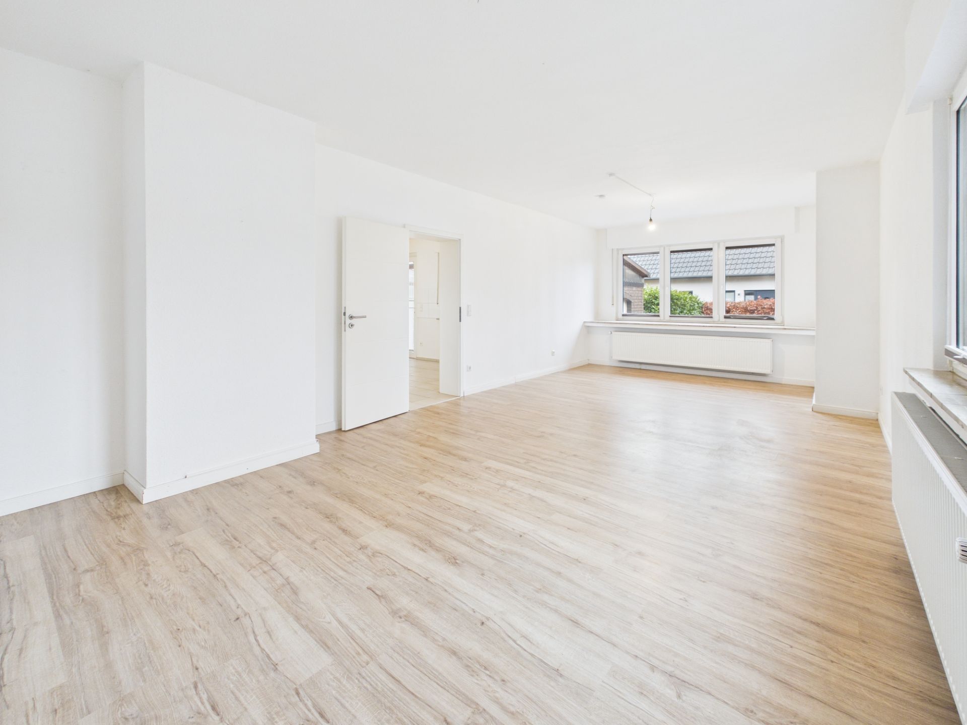 Heller, leerer Raum mit Holzfußboden, weißen Wänden und großen Fenstern.
