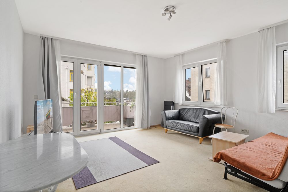 Helles Wohnzimmer mit Sofa, Tisch, Teppich, großen Fenstern und Zugang zum Balkon.