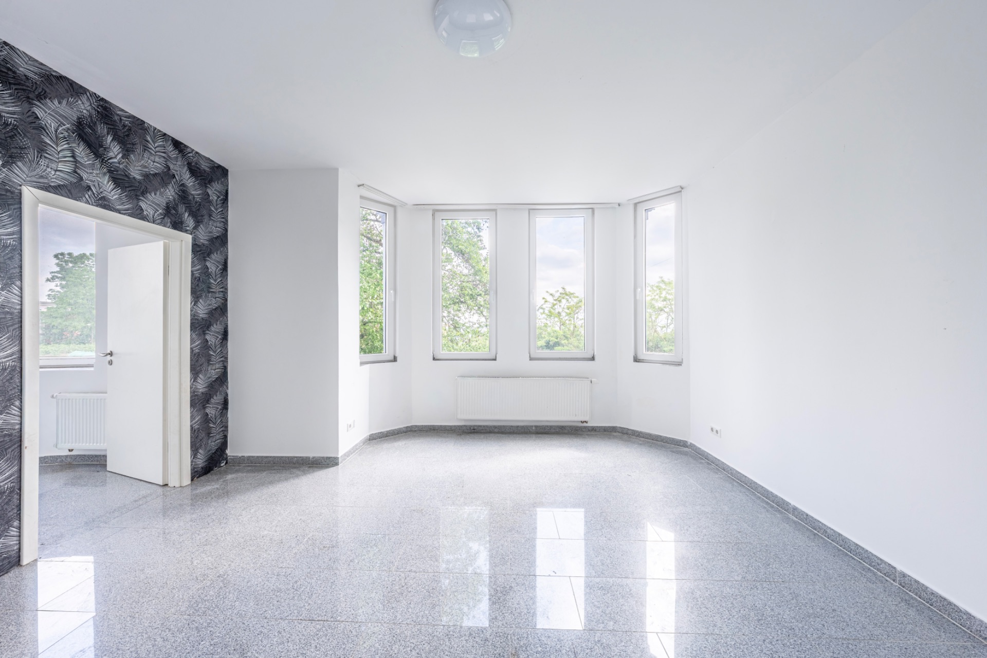 Heller, leerer Raum mit großen Fenstern, grauem Boden und teils gemusterter Wand.