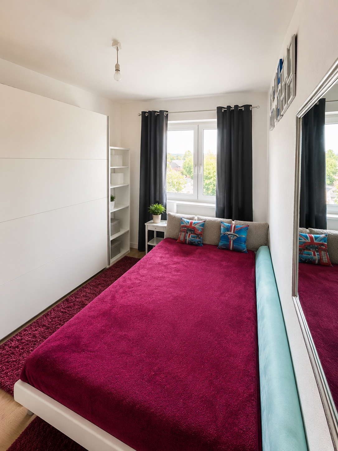 Helles Schlafzimmer mit großem Bett, roter Decke, Kleiderschrank und Fenster mit dunklen Vorhängen.