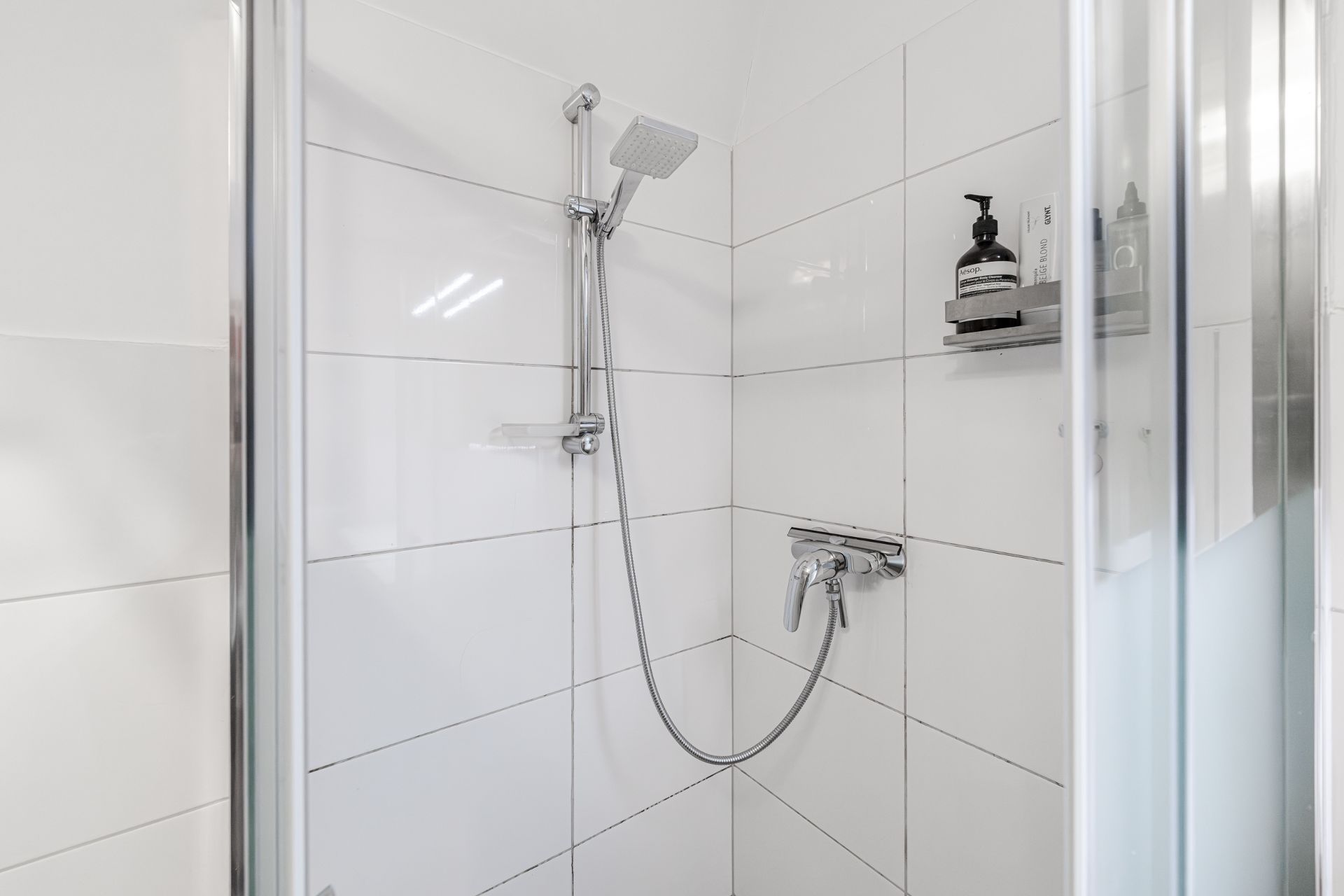 Moderne Dusche mit weiß gefliesten Wänden und Duscharmatur, Regale mit Pflegeprodukten.