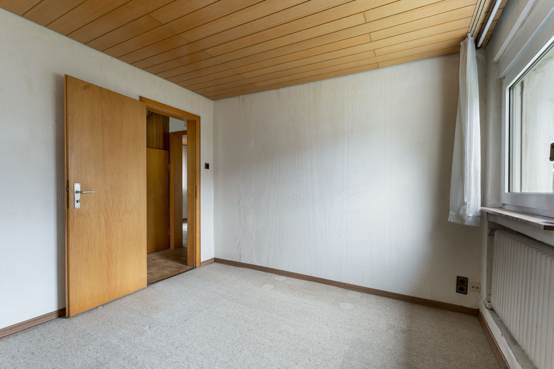 Leeres Zimmer mit Holzdecke, Teppichboden, offenem Holztürrahmen und einem Fenster mit Vorhang.