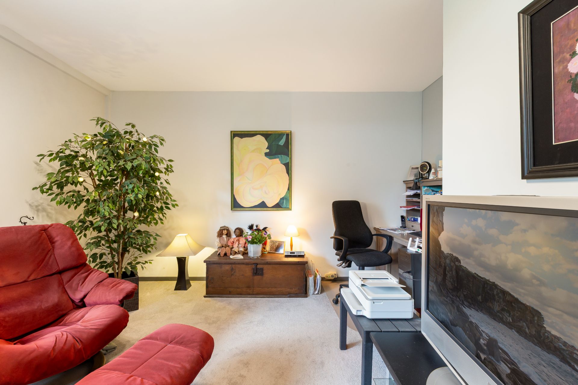 Wohnzimmer mit rotem Sofa, Schreibtisch, grüner Pflanze und Fernseher.