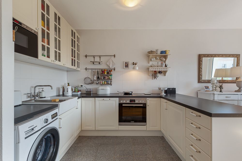Helle Küche mit weißen Schränken, Backofen, Waschmaschine und Wandregalen.