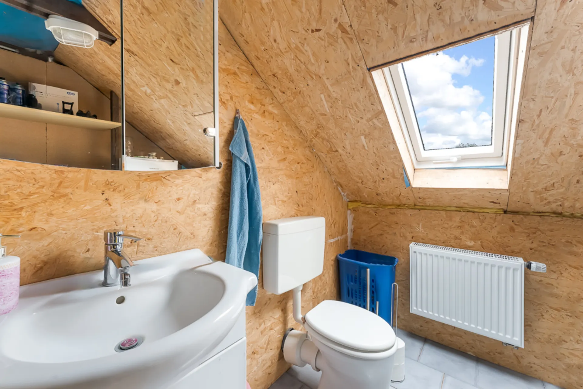 Kleines Badezimmer mit Waschbecken, Toilette, Spiegel, Fenster im Dach und blauer Wäschekorb.