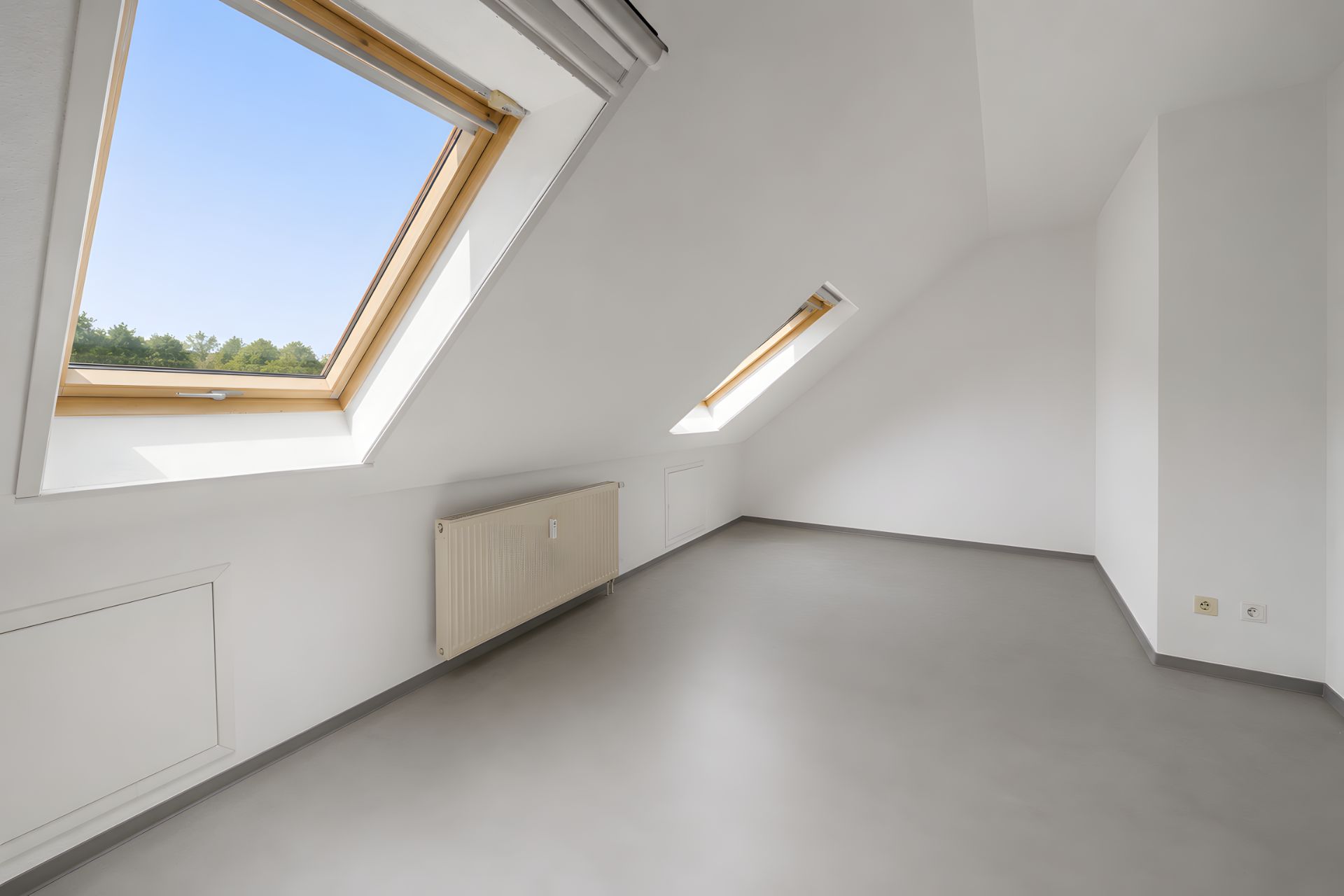 Leerer Dachgeschossraum mit zwei großen Dachfenstern und Heizkörper an der Wand.