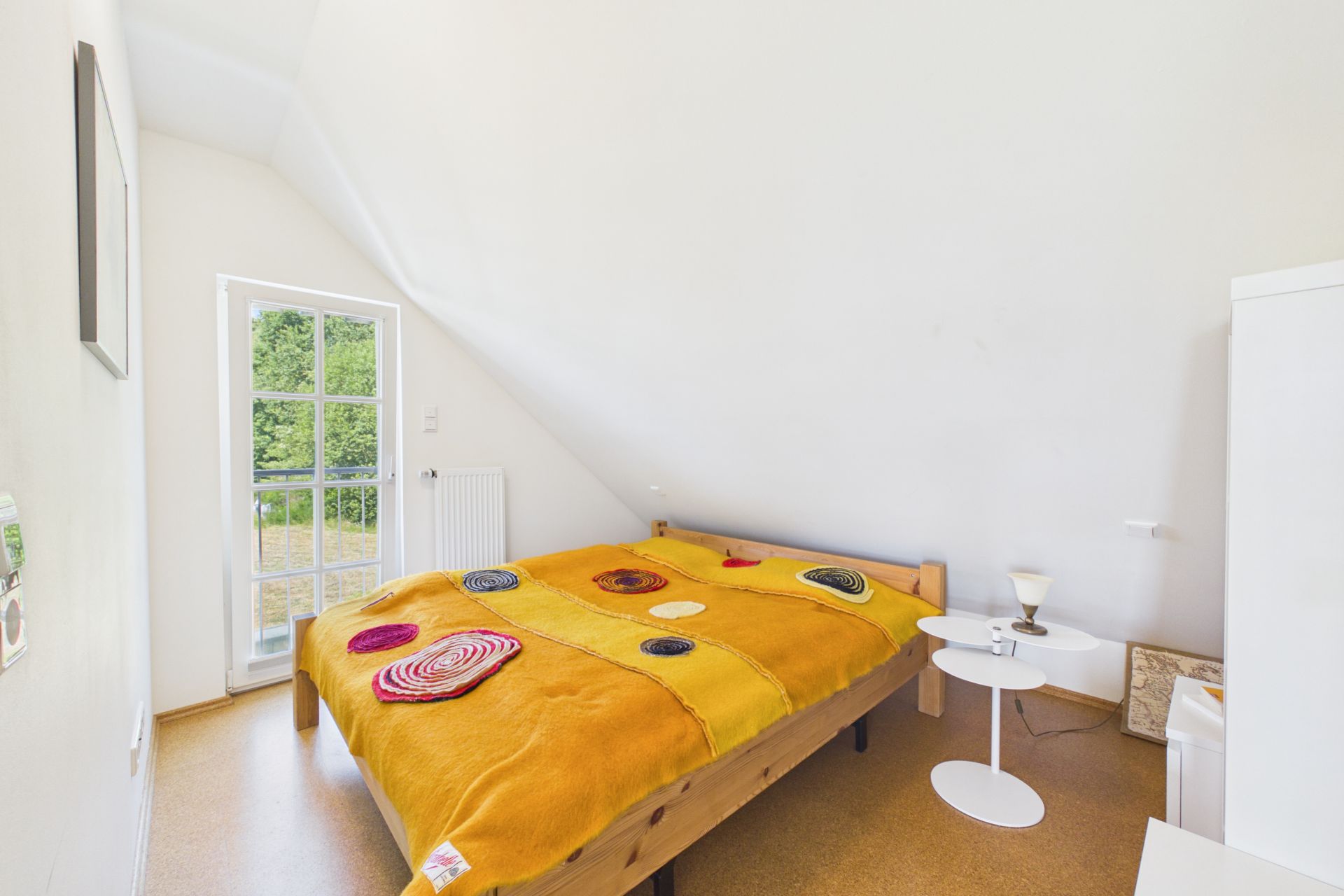 Helles Schlafzimmer mit schräger Decke, Doppelbett mit bunter Decke und kleinem weißen Tisch.
