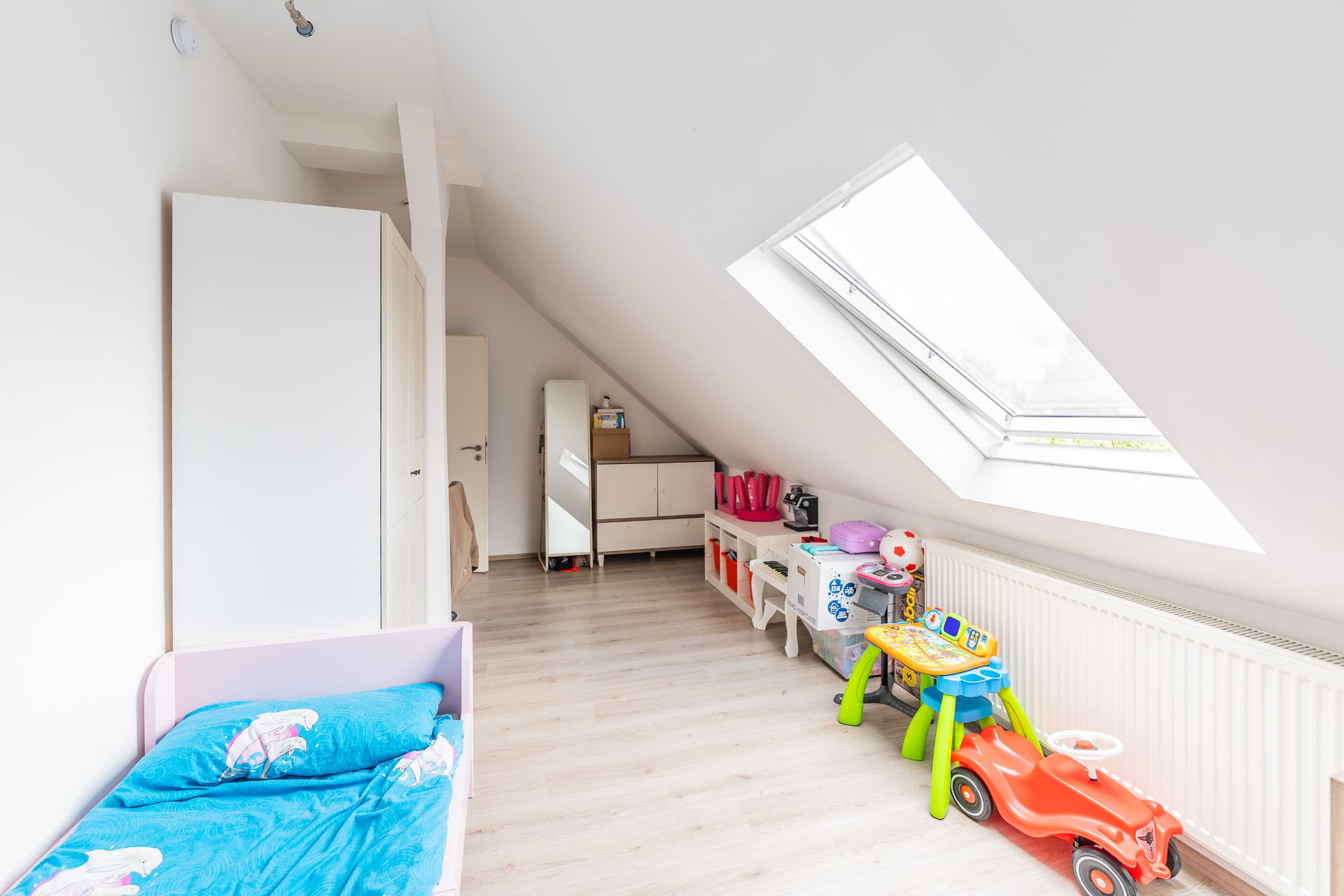 Helles Kinderzimmer mit Schrägdach, Spielzeug, Bett, Schrank und großem Fenster.