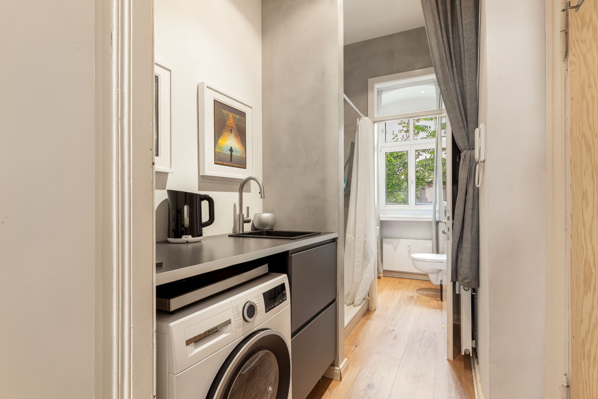Kleines Bad mit Waschmaschine, Waschbecken, Dusche, WC und einem Fenster.