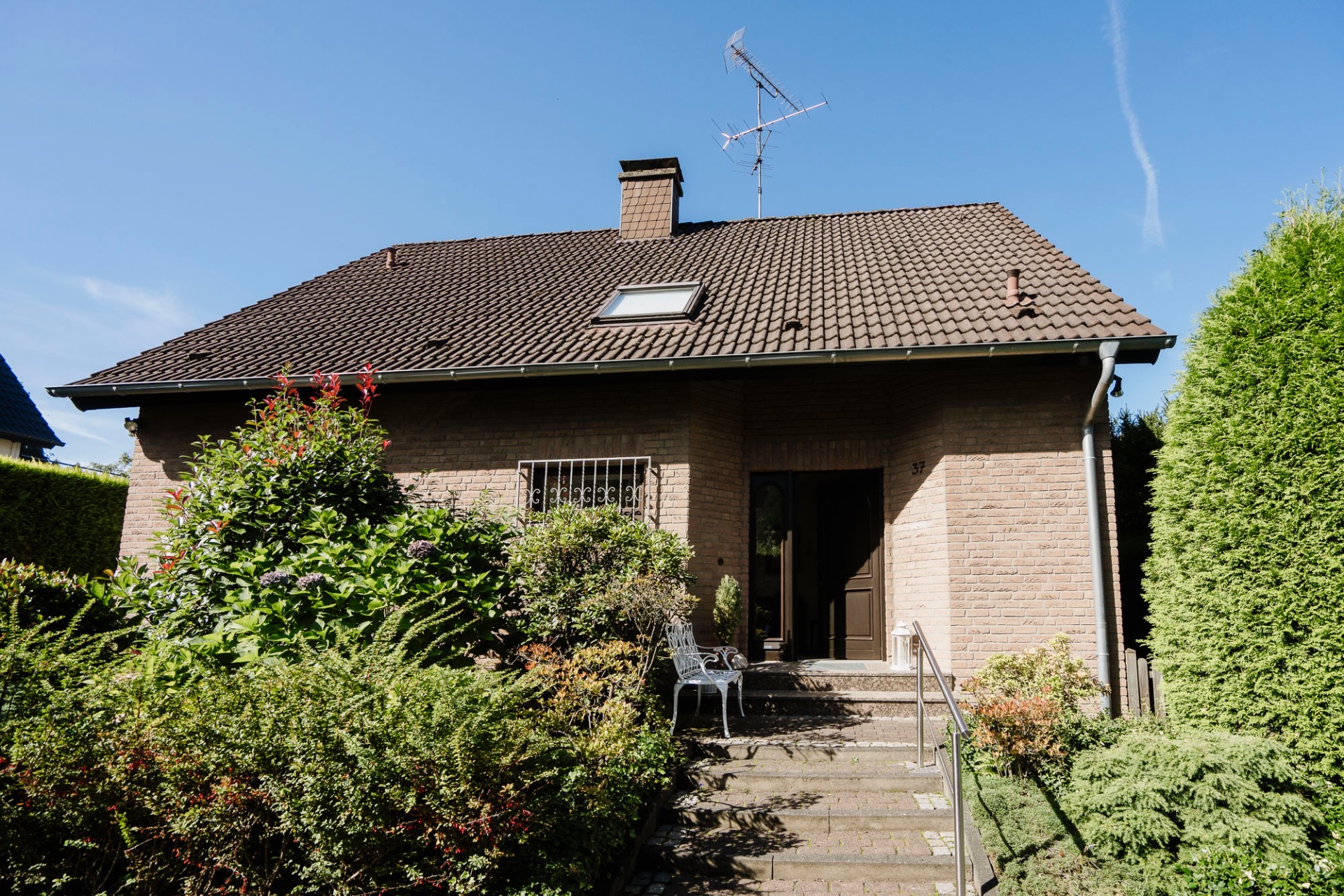 Einfamilienhaus mit dunklem Dach, Garten, Treppe und Gartenmöbeln vor dem Eingang.