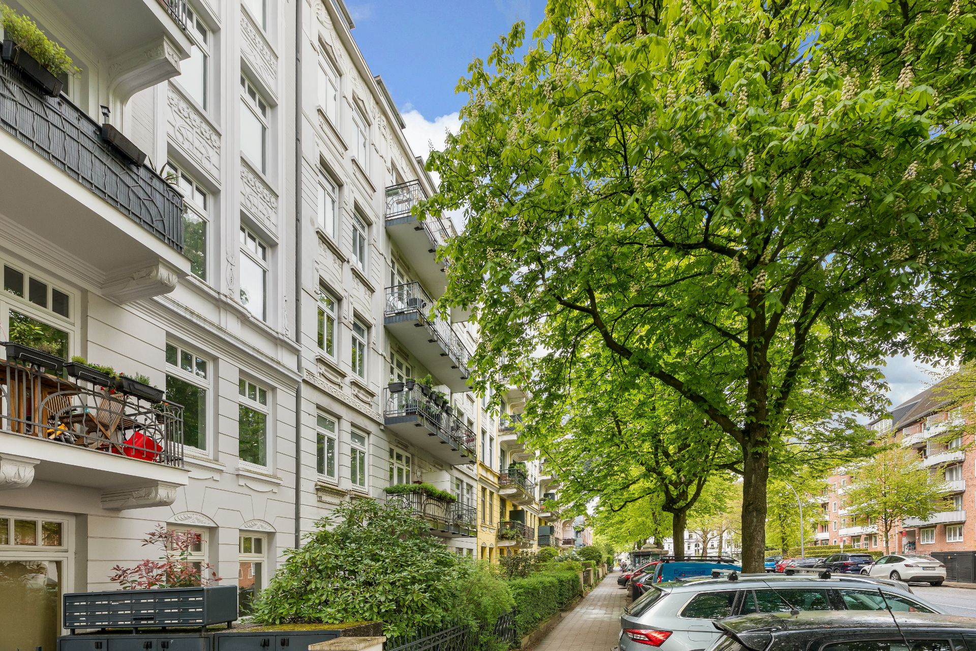 Mehrstöckiges Wohnhaus mit Balkonen an einer von Bäumen gesäumten Straße, geparkte Autos am Straßenrand.