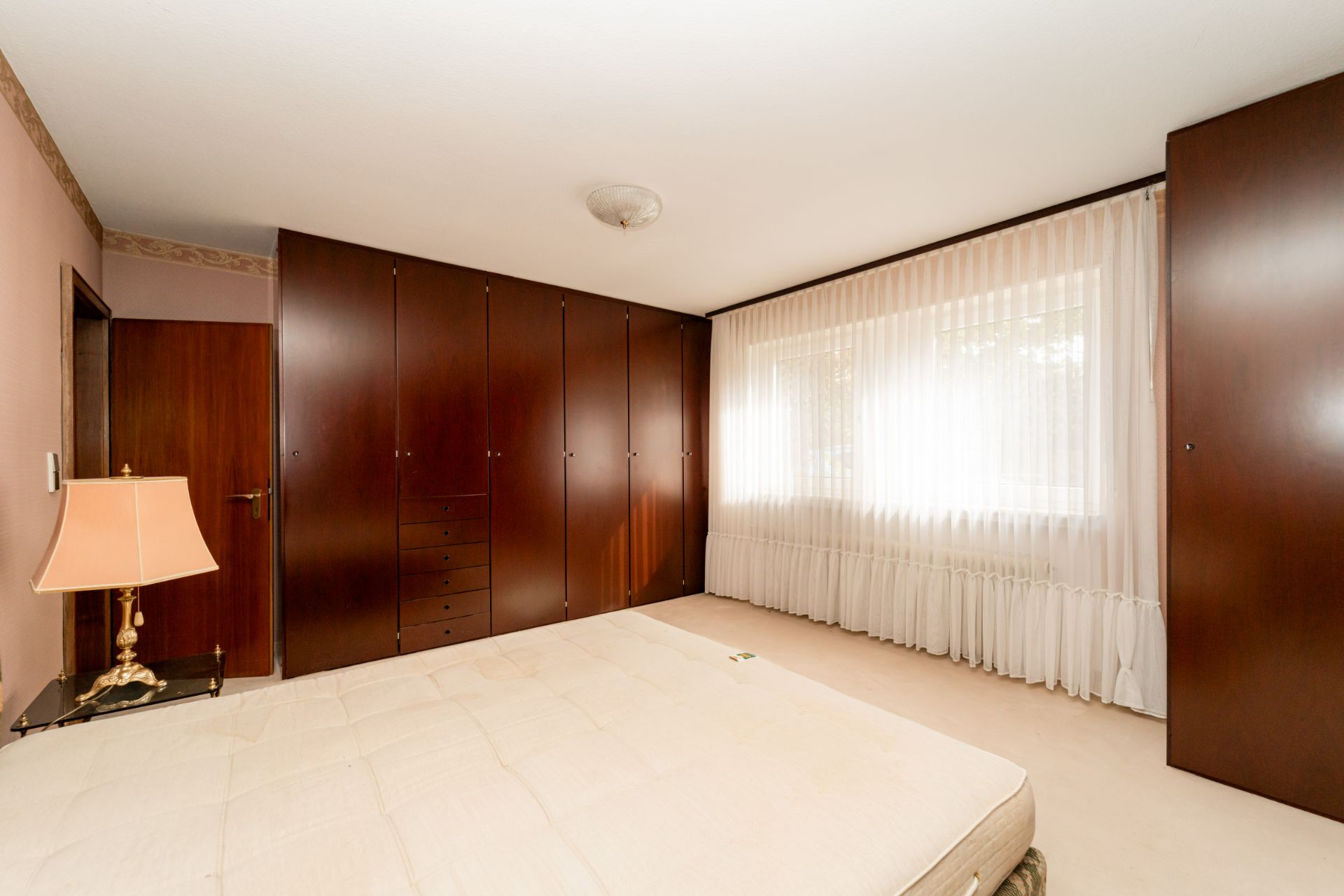Schlafzimmer mit großem Einbauschrank, Fenster mit weißen Vorhängen und Nachttischlampe.