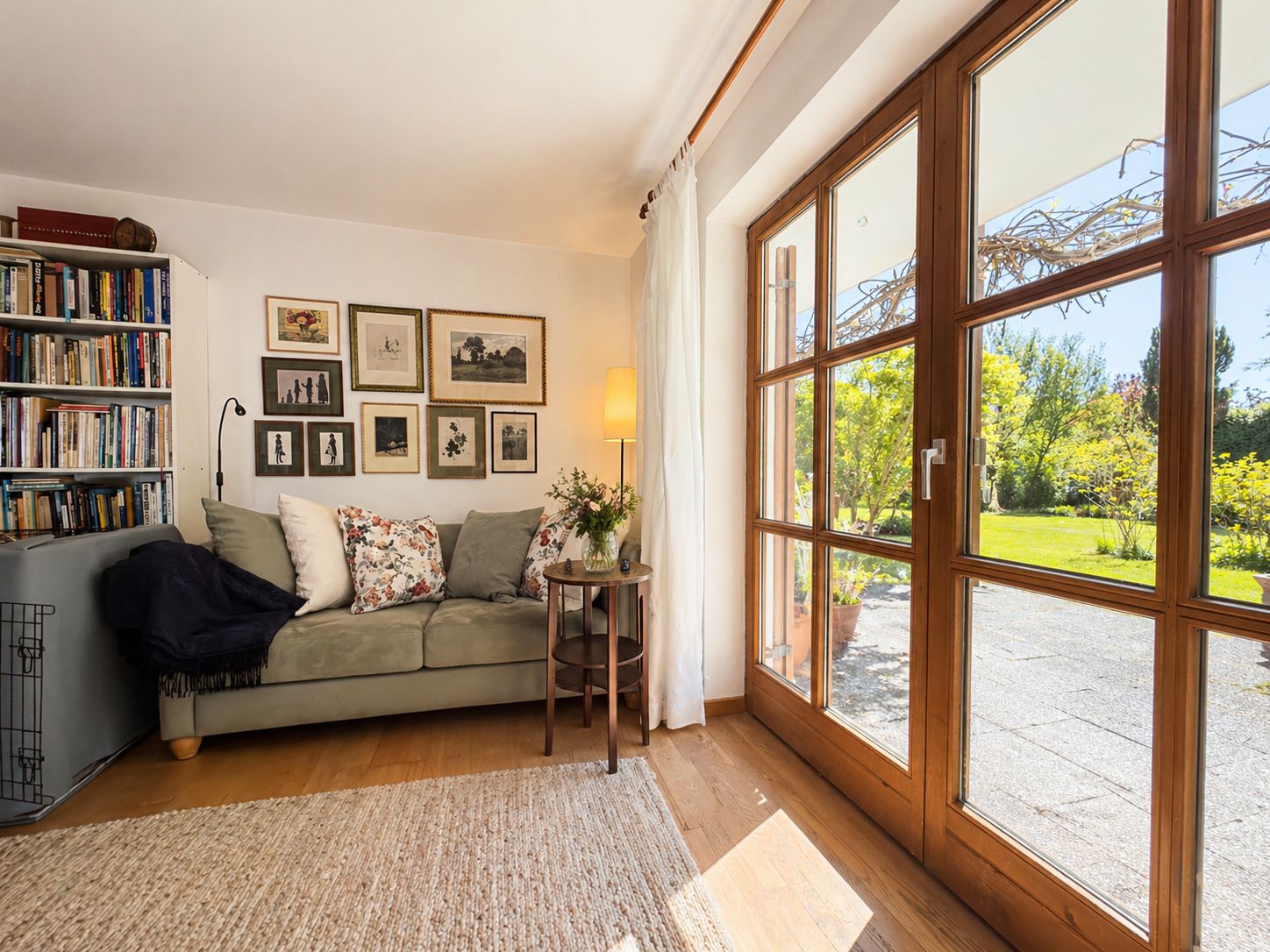 Helles Wohnzimmer mit Sofa, Bücherregal, Bildern an der Wand und großen Fenstern zum Garten.