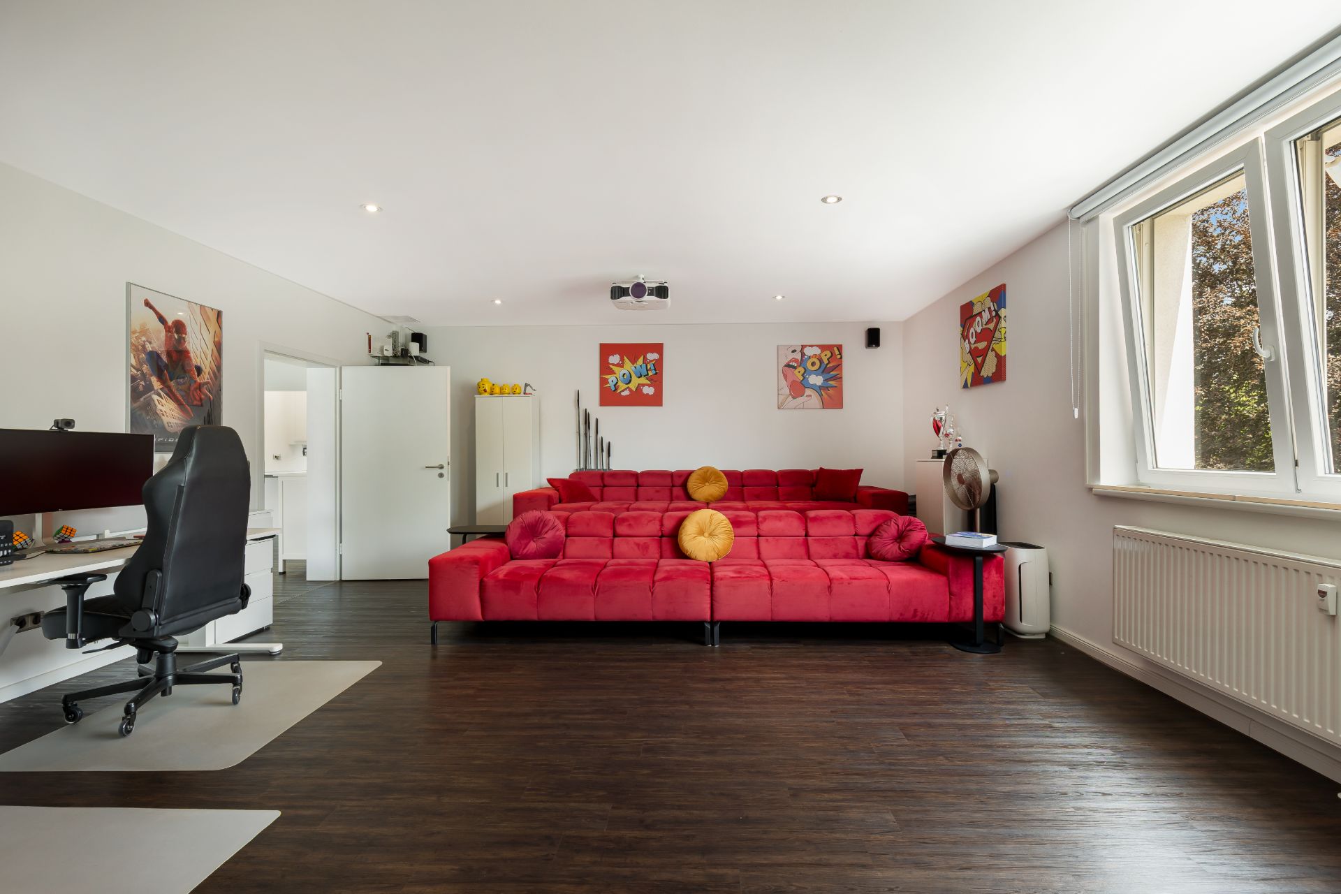 Helles Wohnzimmer mit großer roter Couch, Schreibtisch und mehreren Fenstern.