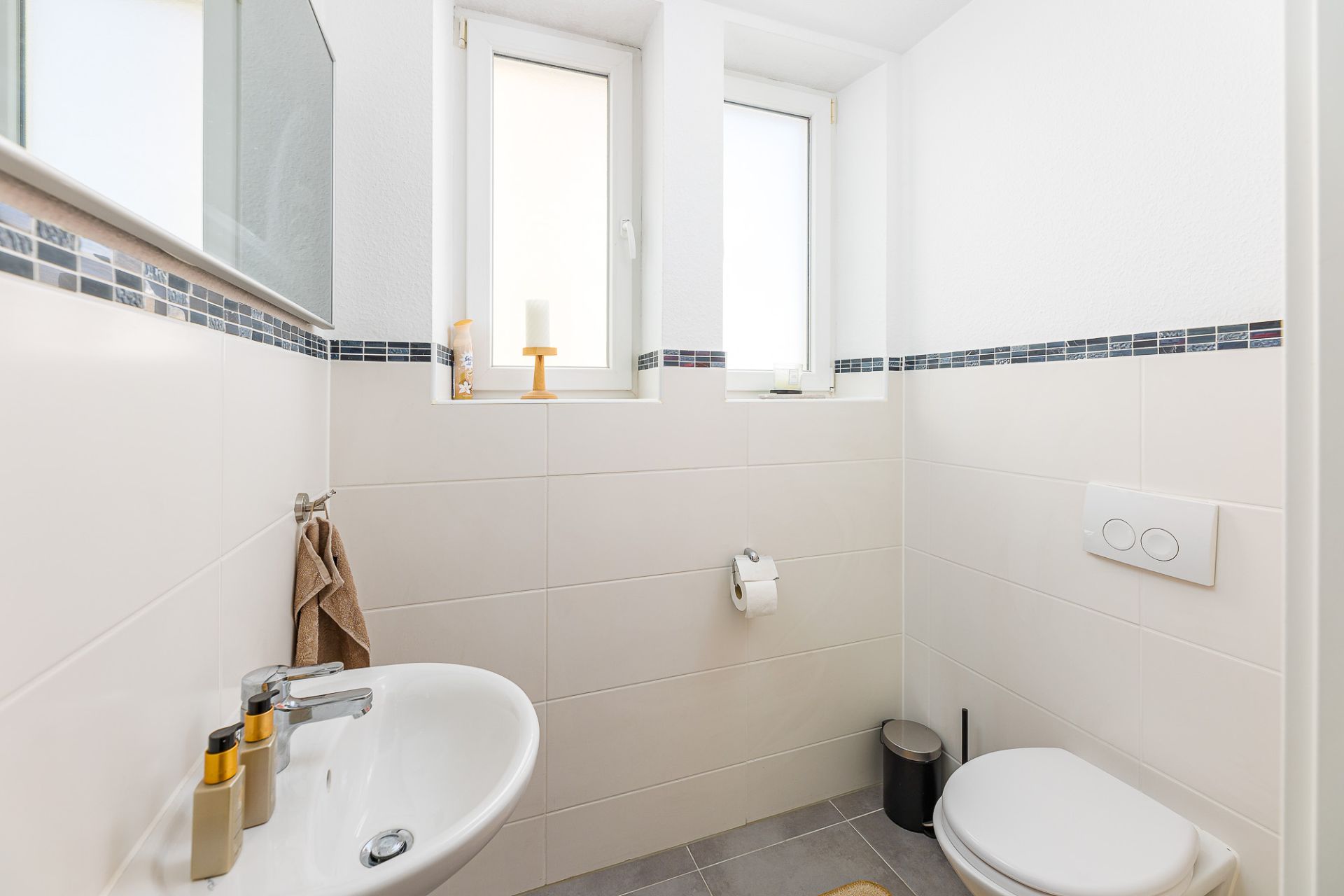 Helles Badezimmer mit Waschbecken, Wandtoilette, zwei Fenstern und Handtuchhalter.