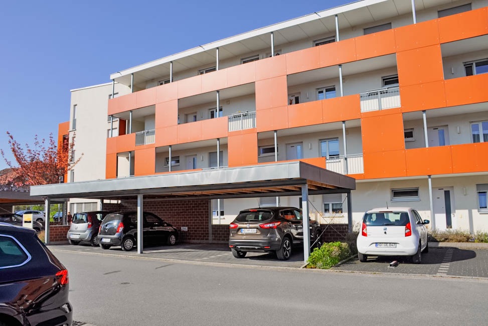 Mehrfamilienhaus mit oranger Fassade und überdachten Parkplätzen davor.