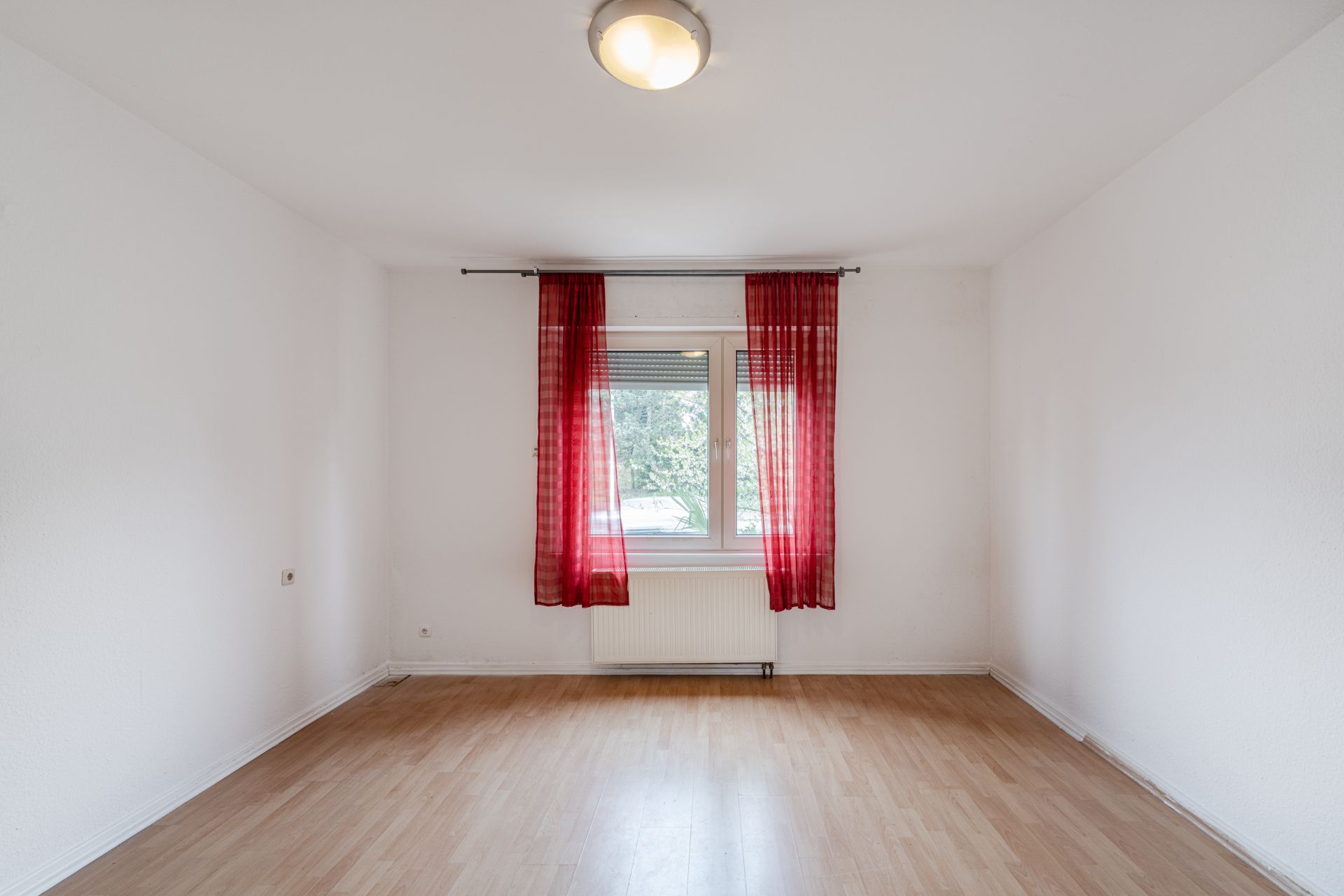 Leeres Zimmer mit hellem Boden, weißer Wand und Fenster mit roten Vorhängen.