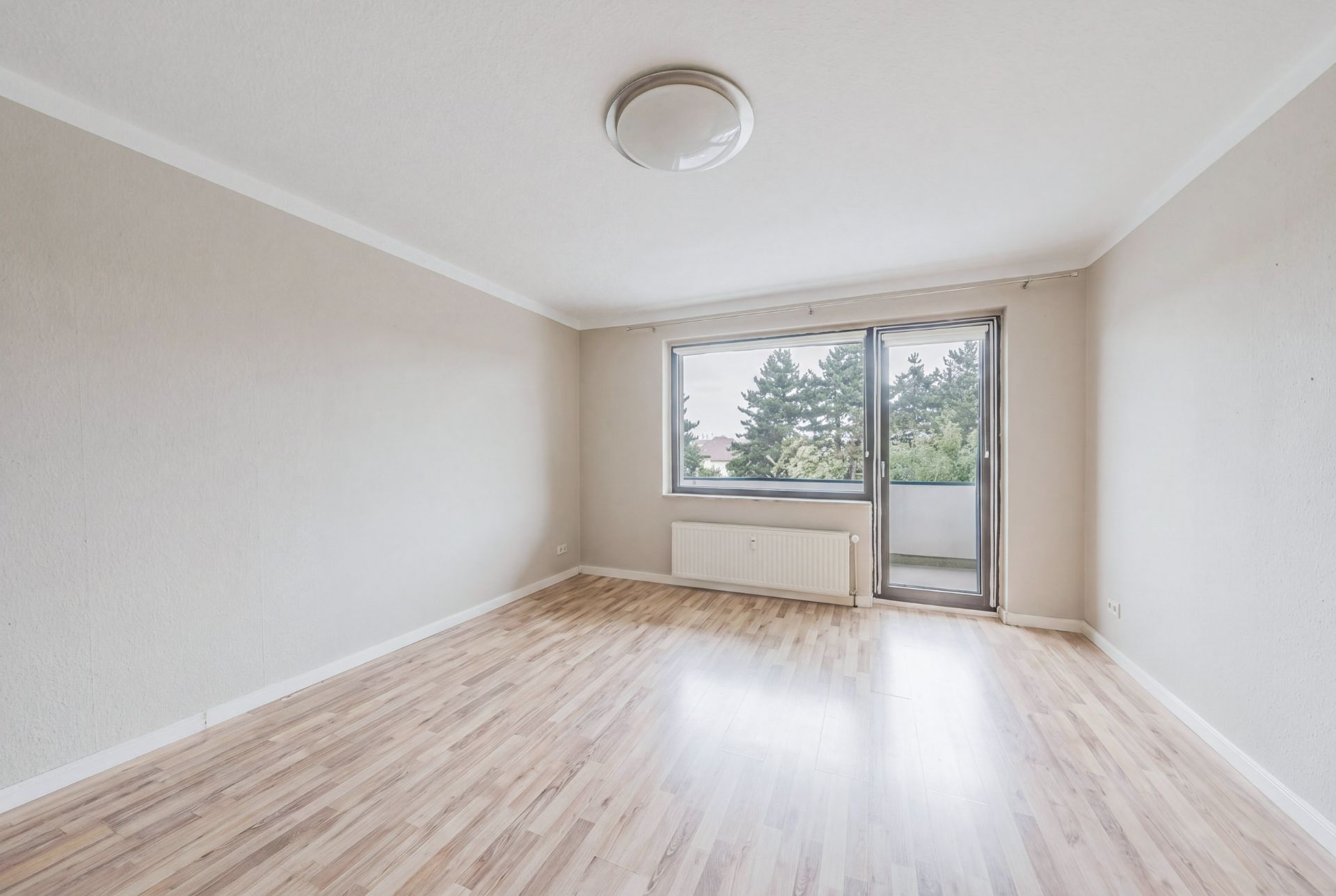 Leeres, helles Zimmer mit Holzfußboden, Fenster und Tür zum Balkon, Blick auf Bäume.