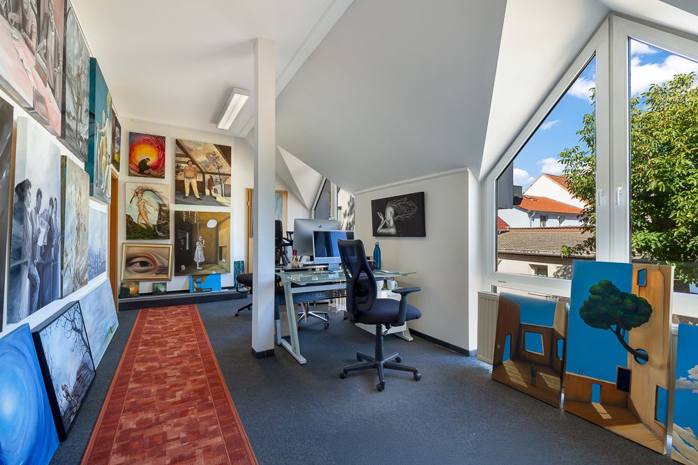 Helles Arbeitszimmer mit Schreibtischen, Bürostühlen, vielen Gemälden und großem Fenster.
