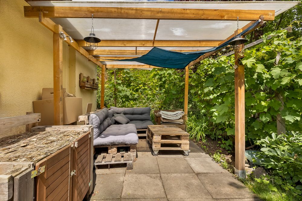Überdachte Terrasse mit Holzgestell, Ecksofa, Palettenmöbeln und viel Grün im Hintergrund.