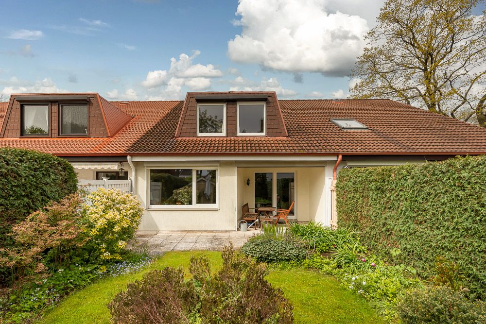 Rückseite eines Hauses mit Terrasse, Garten und großen Fenstern, umgeben von Hecken und Pflanzen.