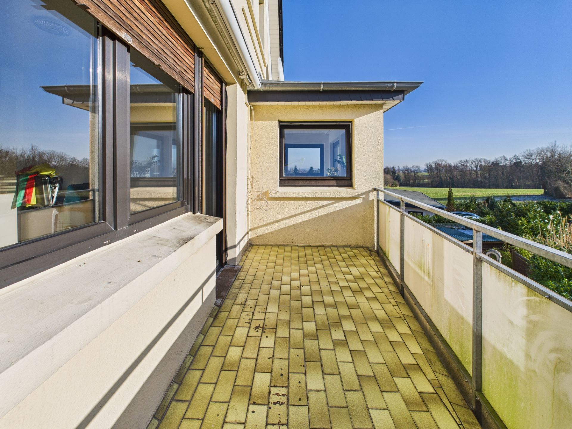 Geräumiger Balkon mit gelben Fliesen und Geländer, Blick auf Garten und Felder.
