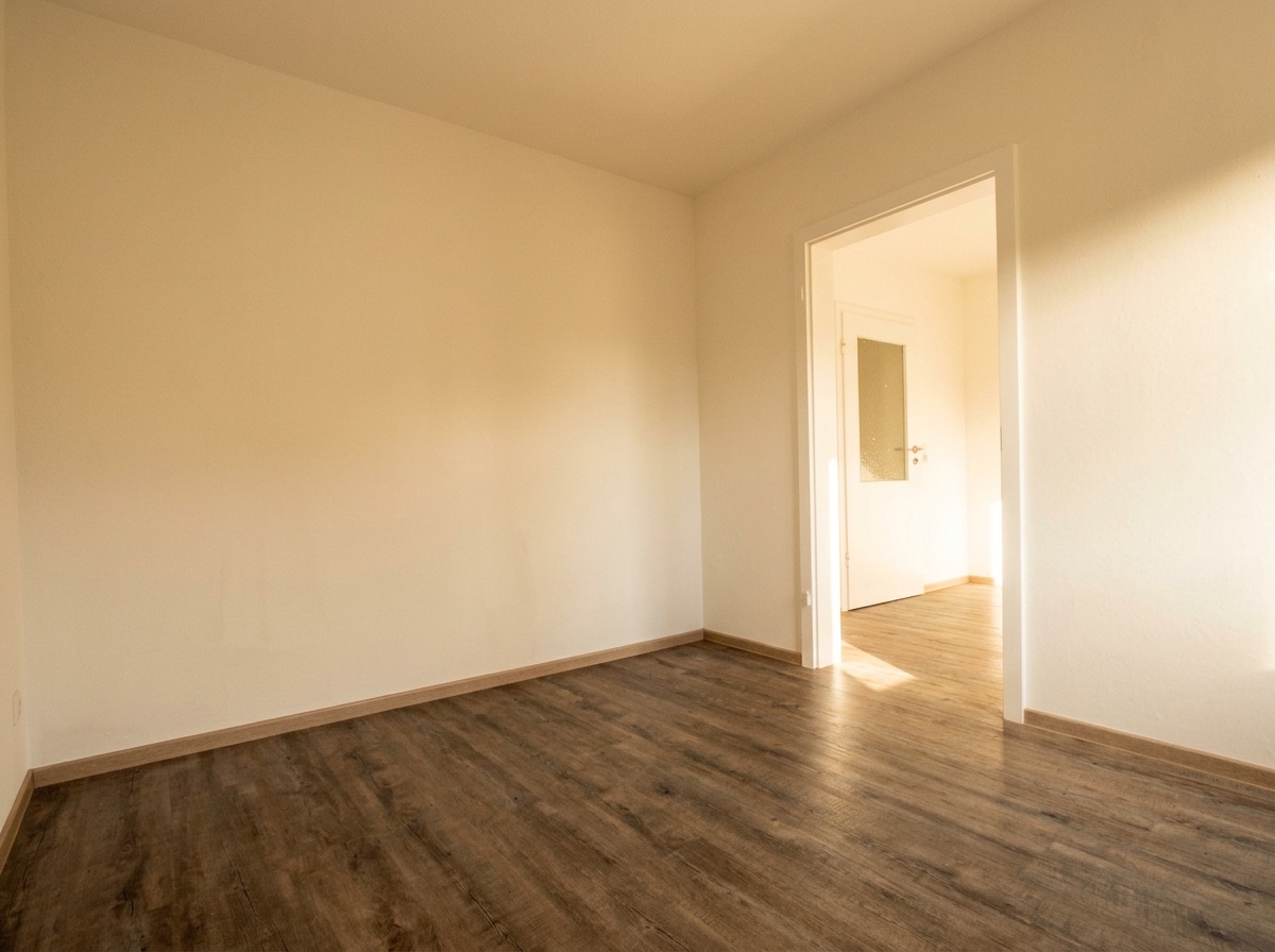 Leerer Raum mit hellem Holzboden und weißer Wand, offene Tür führt in einen weiteren Raum.