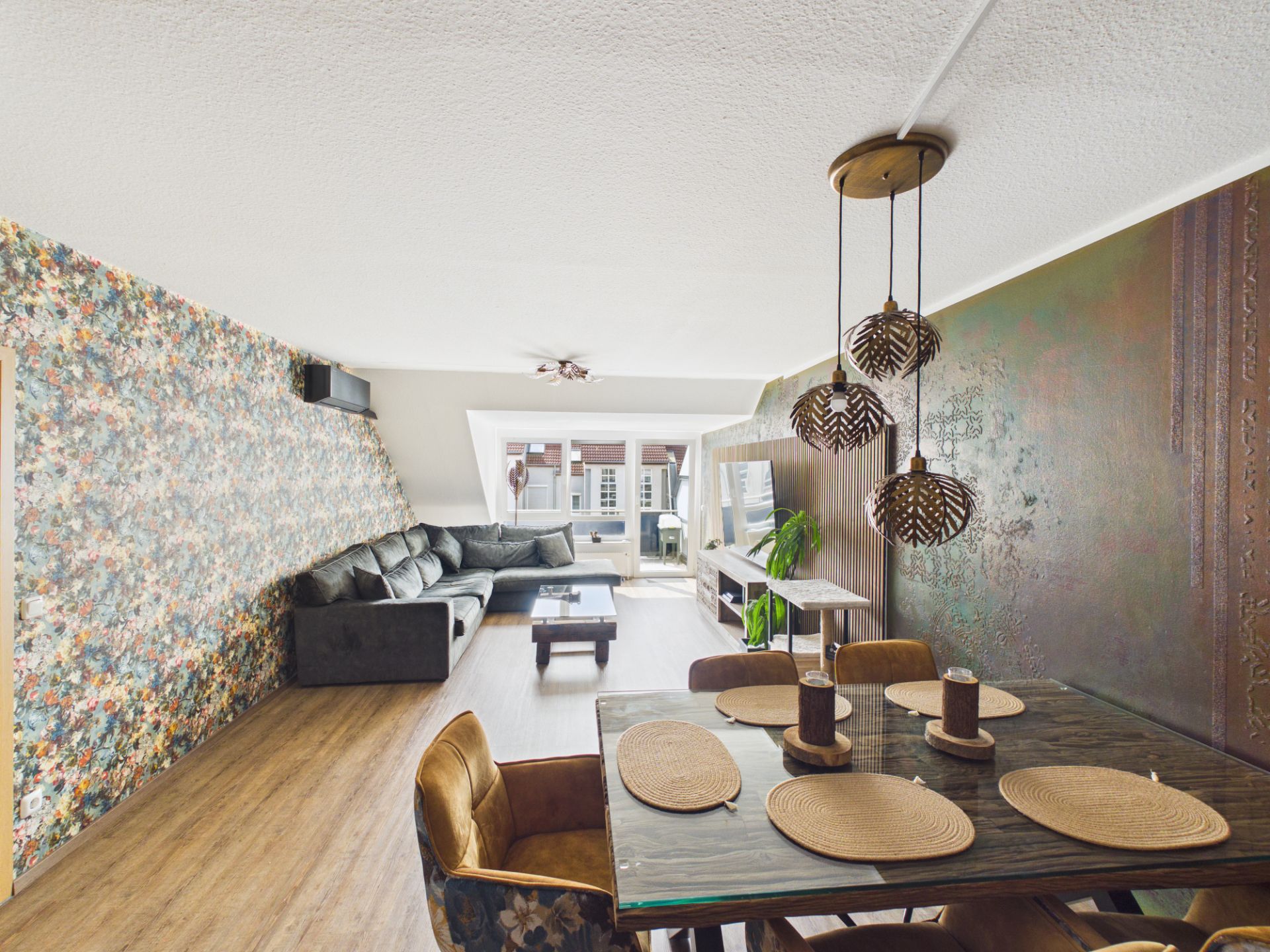 Helles Wohnzimmer mit Ecksofa, Esstisch und bunter Tapete, Holzboden und großen Fenstern.