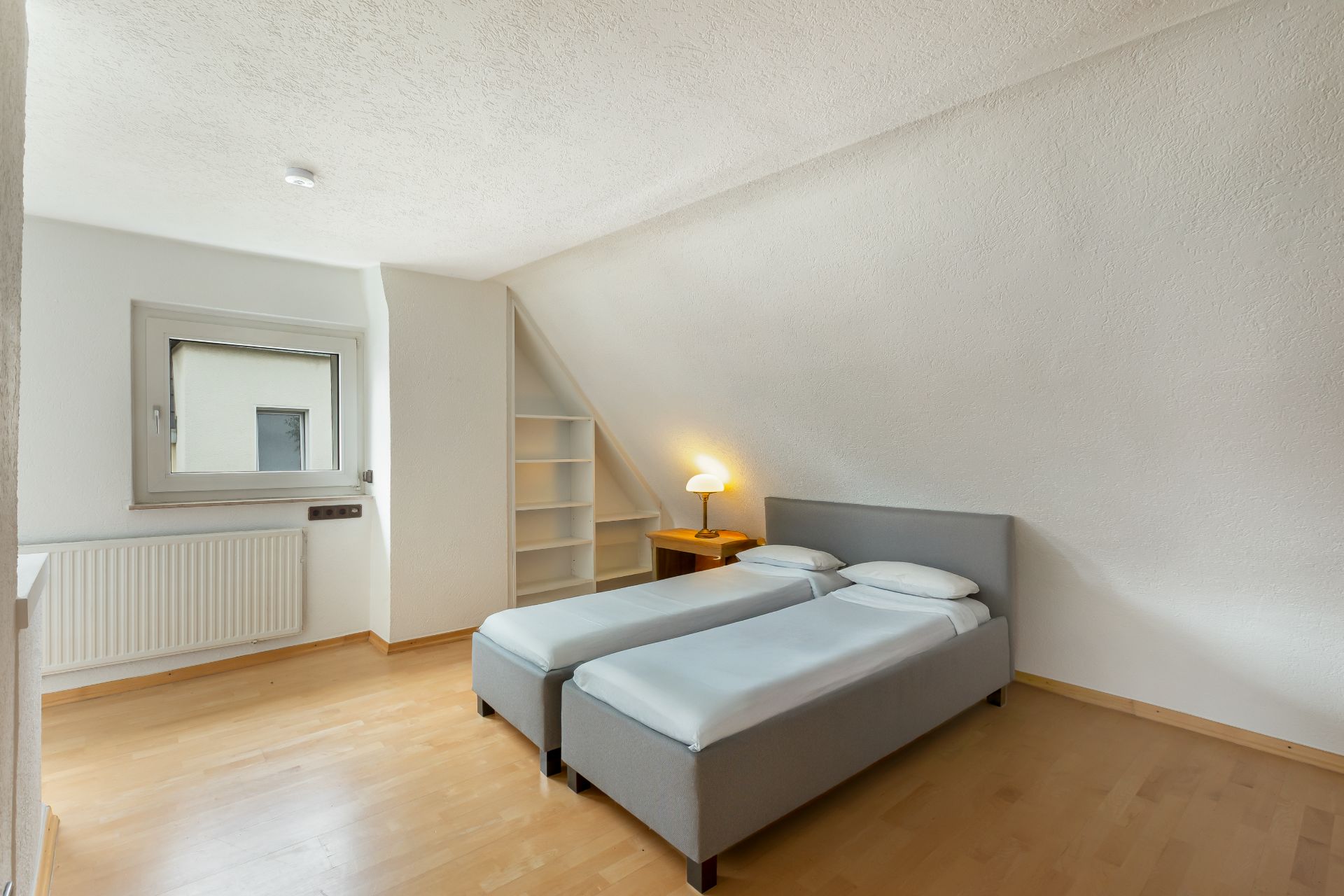 Helles Schlafzimmer mit zwei Einzelbetten, Nachttisch, Lampe und Regal unter der Dachschräge.