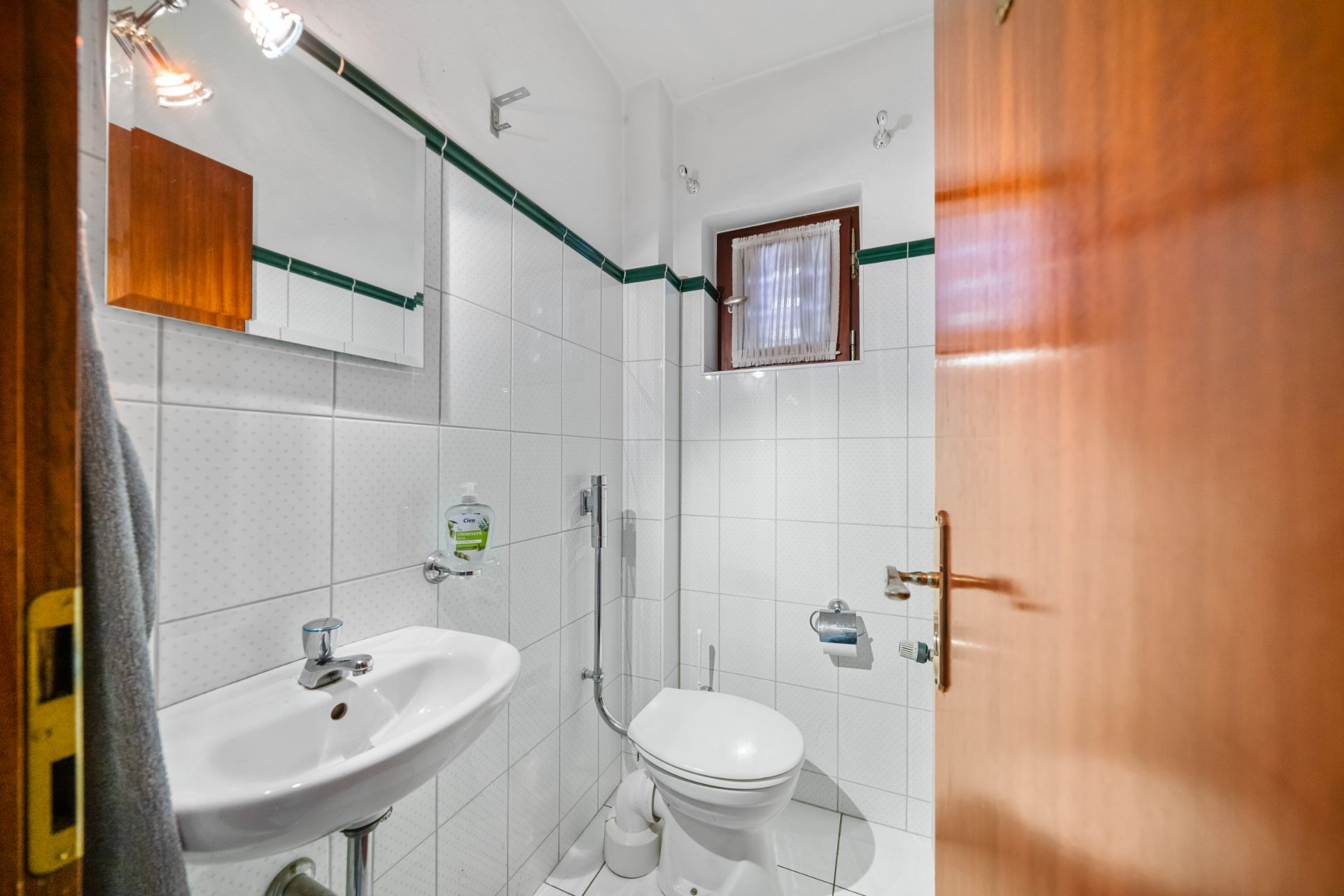 Kleines Badezimmer mit Waschbecken, WC, Spiegel und kleinem Fenster.
