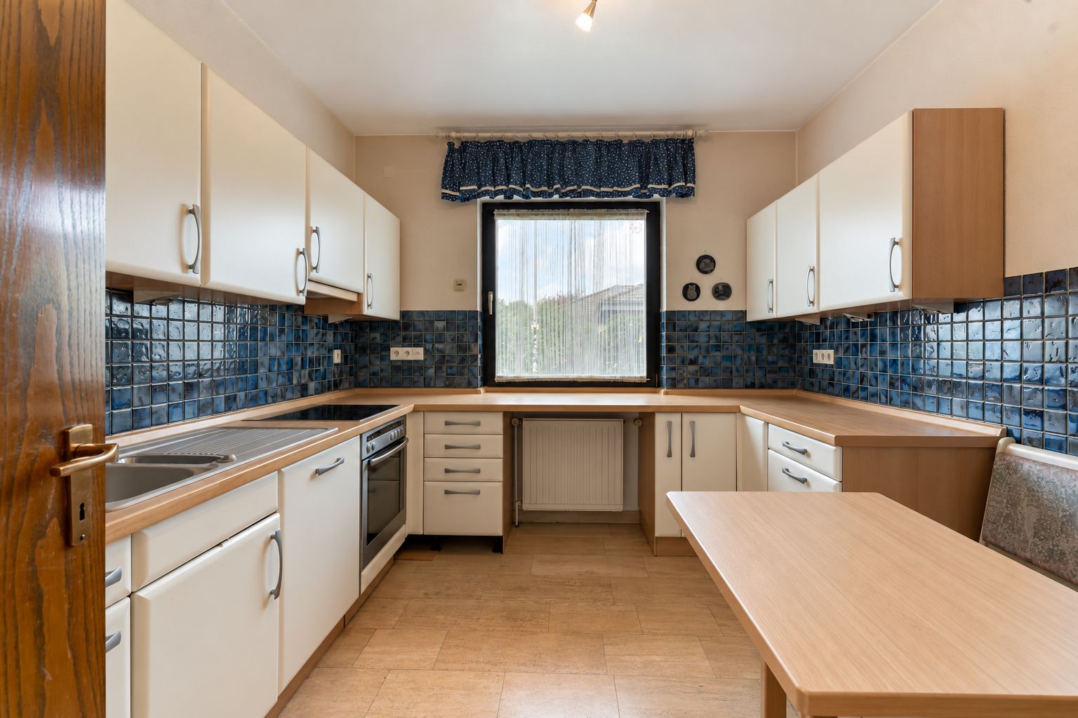 Küche mit weißen Schränken, blau gekachelter Wand, großem Fenster und Holztisch.