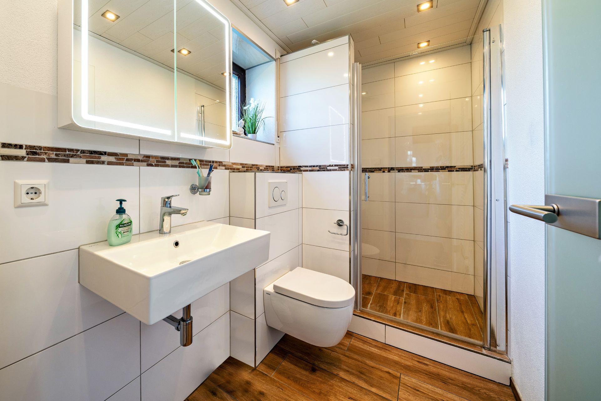 Modernes Badezimmer mit Waschbecken, Wandtoilette, beleuchtetem Spiegel und Dusche mit Glastür.