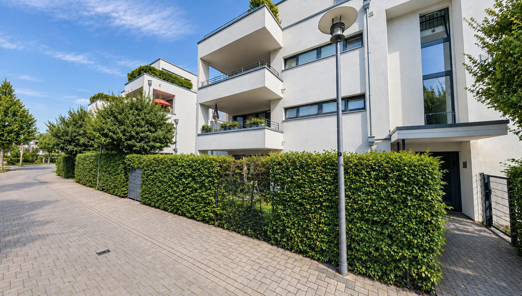 Moderne Wohnanlage mit Balkonen und grüner Hecke an einer gepflasterten Straße.