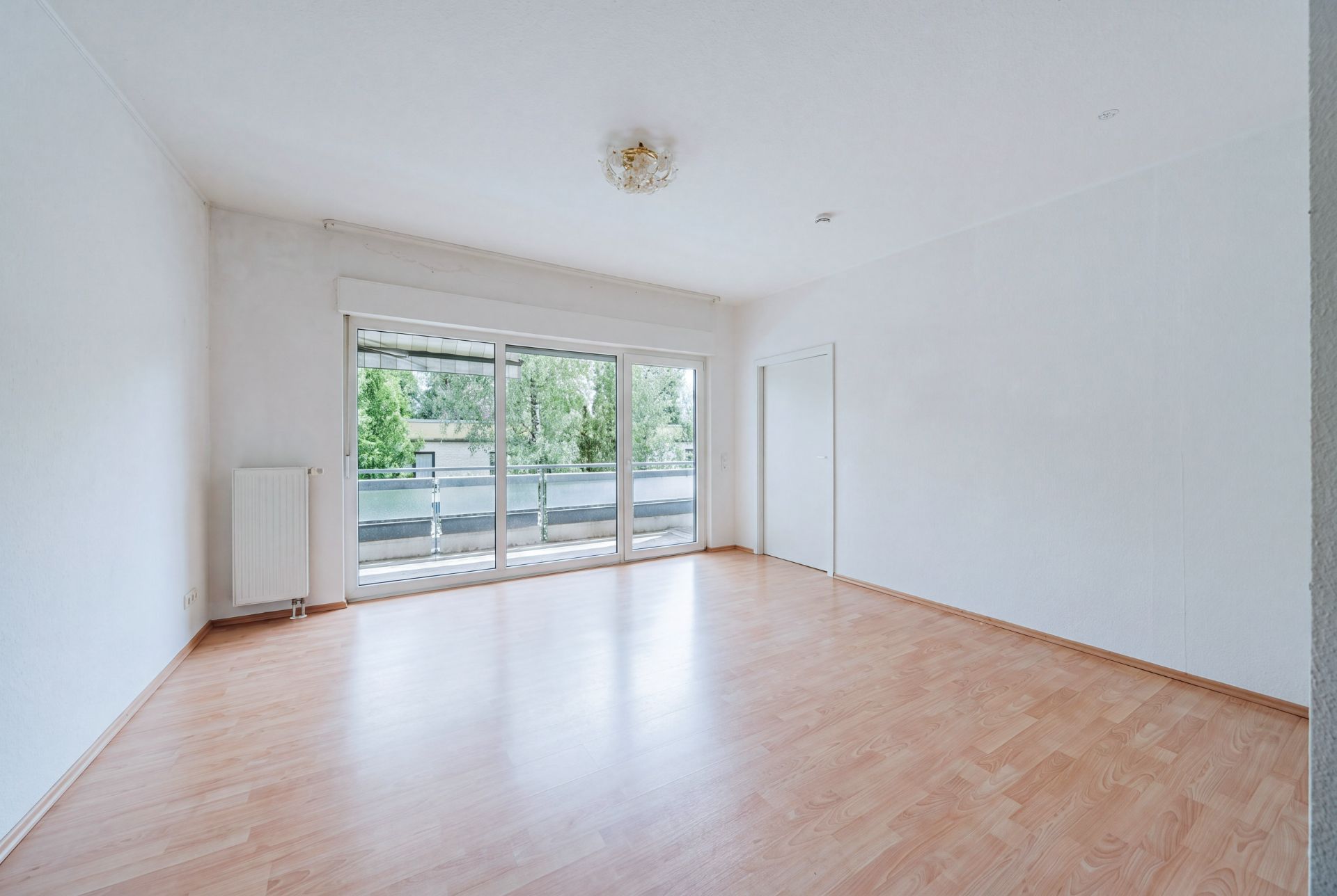 Helles, leeres Zimmer mit Holzfußboden und großen Fenstern zu einem Balkon.