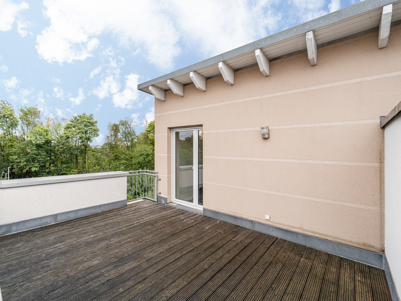 Große Terrasse mit Holzboden, einer Glastür und Blick auf grüne Bäume.